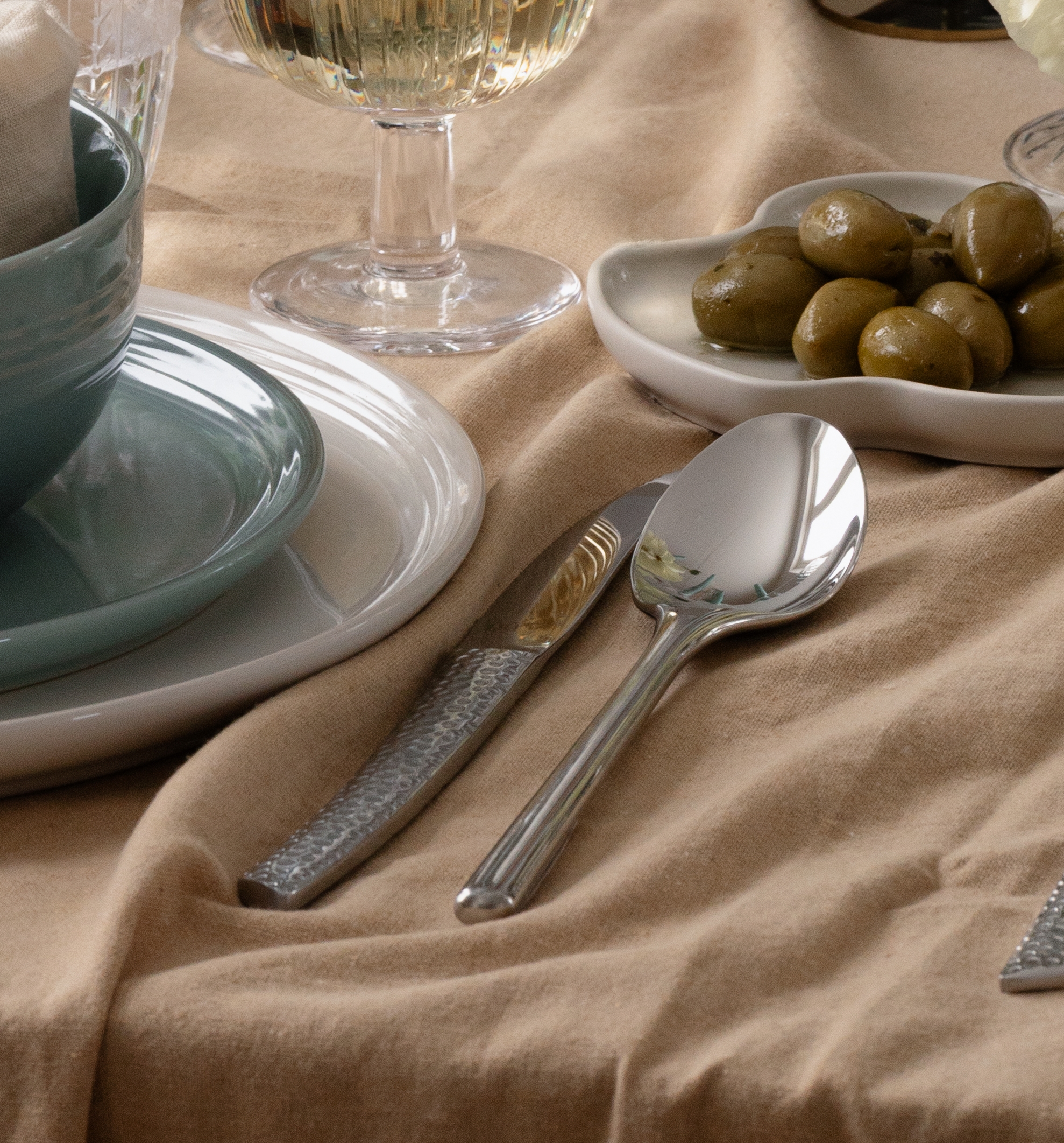 Henkilön käsi pitelee viinilasia ruokapöydällä, jolla on vaaleansinisiä kynttilöitä, valkoisia kukkia, sinivihreät astiat ja ruskea pöytäliina.