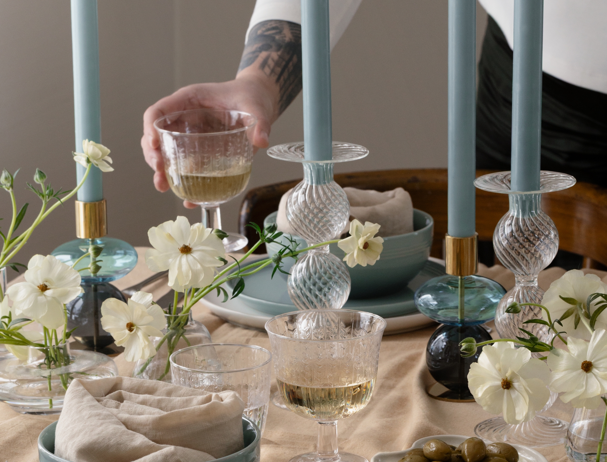 Tatuoitu käsivarsi asettaa viinilasin ruokapöydälle, jolla on sinisiä kynttilöitä, valkoisia kukkia, sinivihreät ja valkoiset astiat, leipää ja oliiveja vaaleanruskealla liinalla.