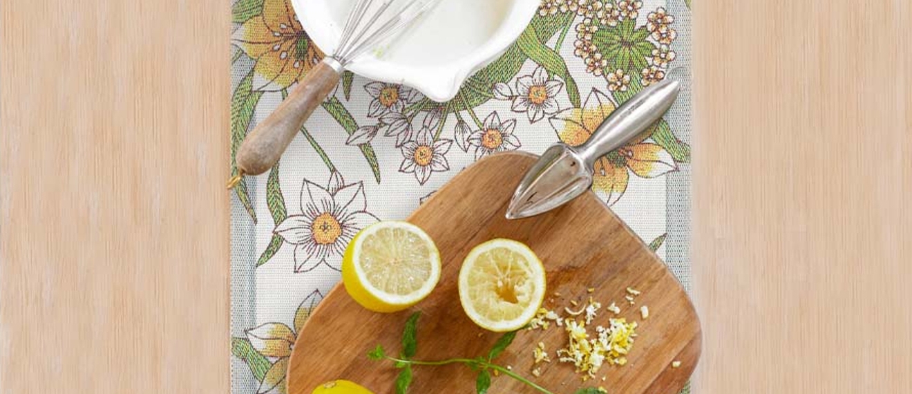 Kukallinen kaitaliina jonka päällä puinen leikkuulauta sitruunoiden kera sekä taikinakulho vispilän kera