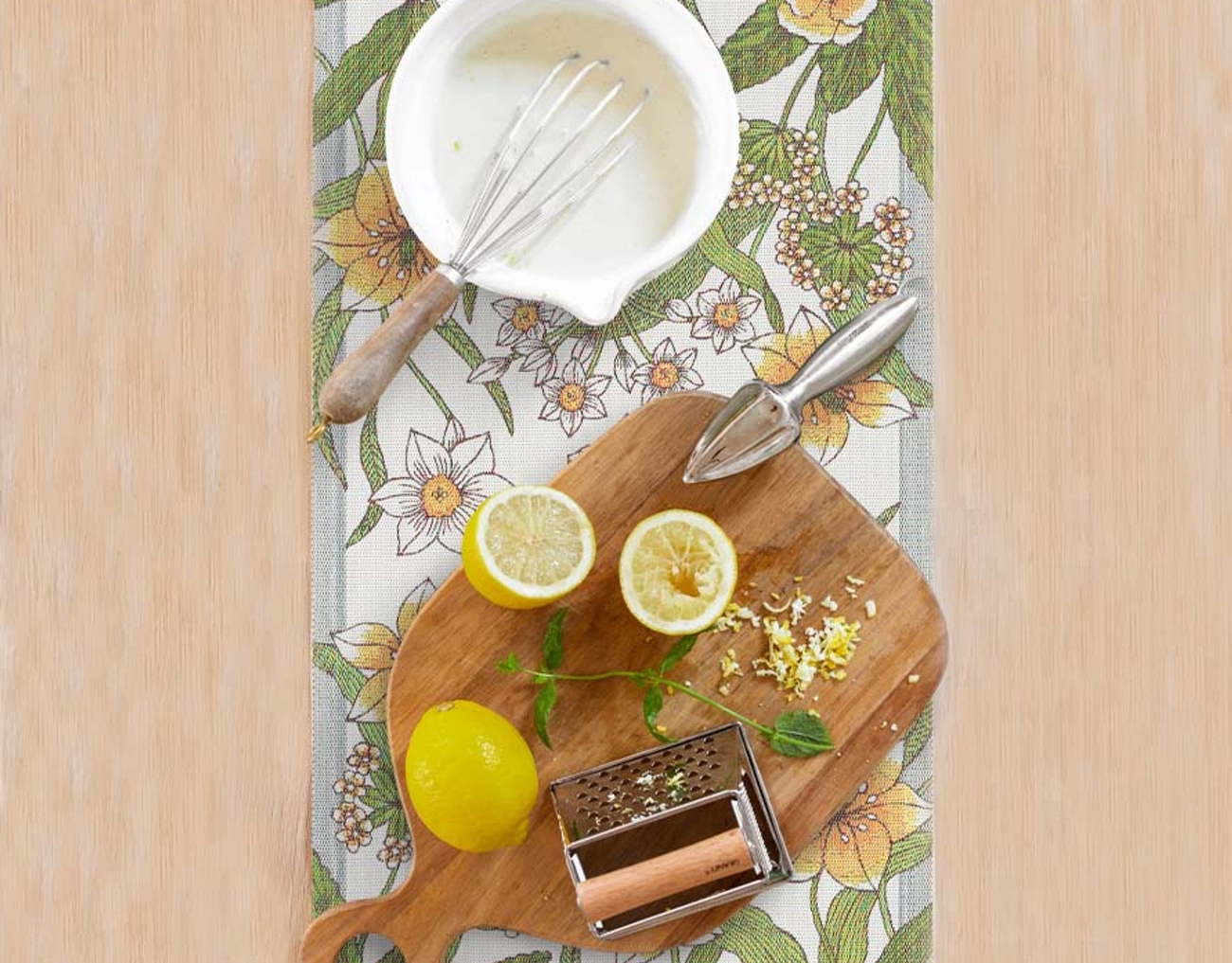 Kukallinen kaitaliina jonka päällä puinen leikkuulauta sitruunoiden kera sekä taikinakulho vispilän kera