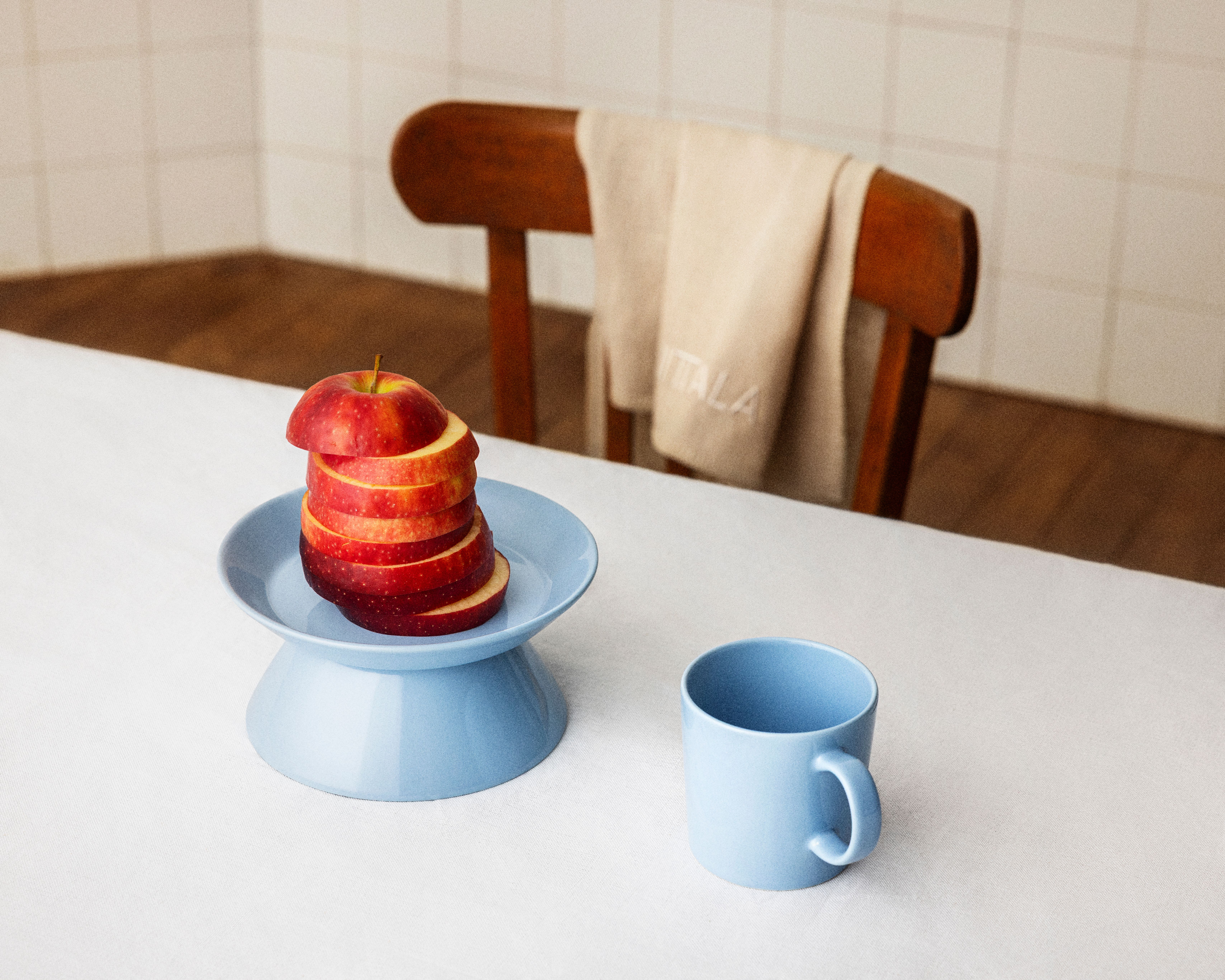 Pino viipaloituja punaisia ​​omenoita vaaleansinisellä jalustalautasella ja yhteensopiva muki valkoisella pöydällä. Taustalla puinen tuoli ja beigenvärinen pyyhe.