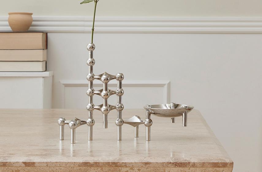 Minimalistinen sisustusasetelma, jossa metallinen modulaarinen kynttilänjalka ja maljakko seisovat vaalealla kivipöydällä neutraalin seinän edessä.