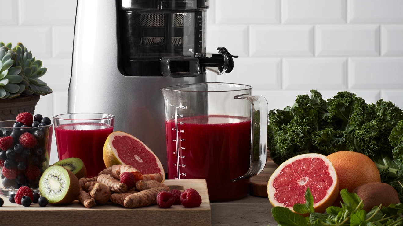 Hurom juicepress med röd juice samt grapefrukt, kiwi och hallon på skärbräda framför