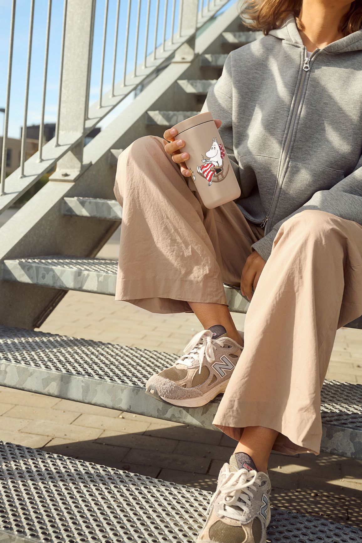 Person sitter avslappnat på en utomhustrappa i beige byxor och sneakers och håller en beige mugg med Mumin-motiv.