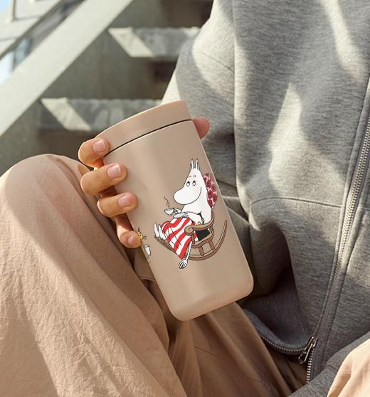 Person sitter avslappnat på en utomhustrappa i beige byxor och sneakers och håller en beige mugg med Mumin-motiv.