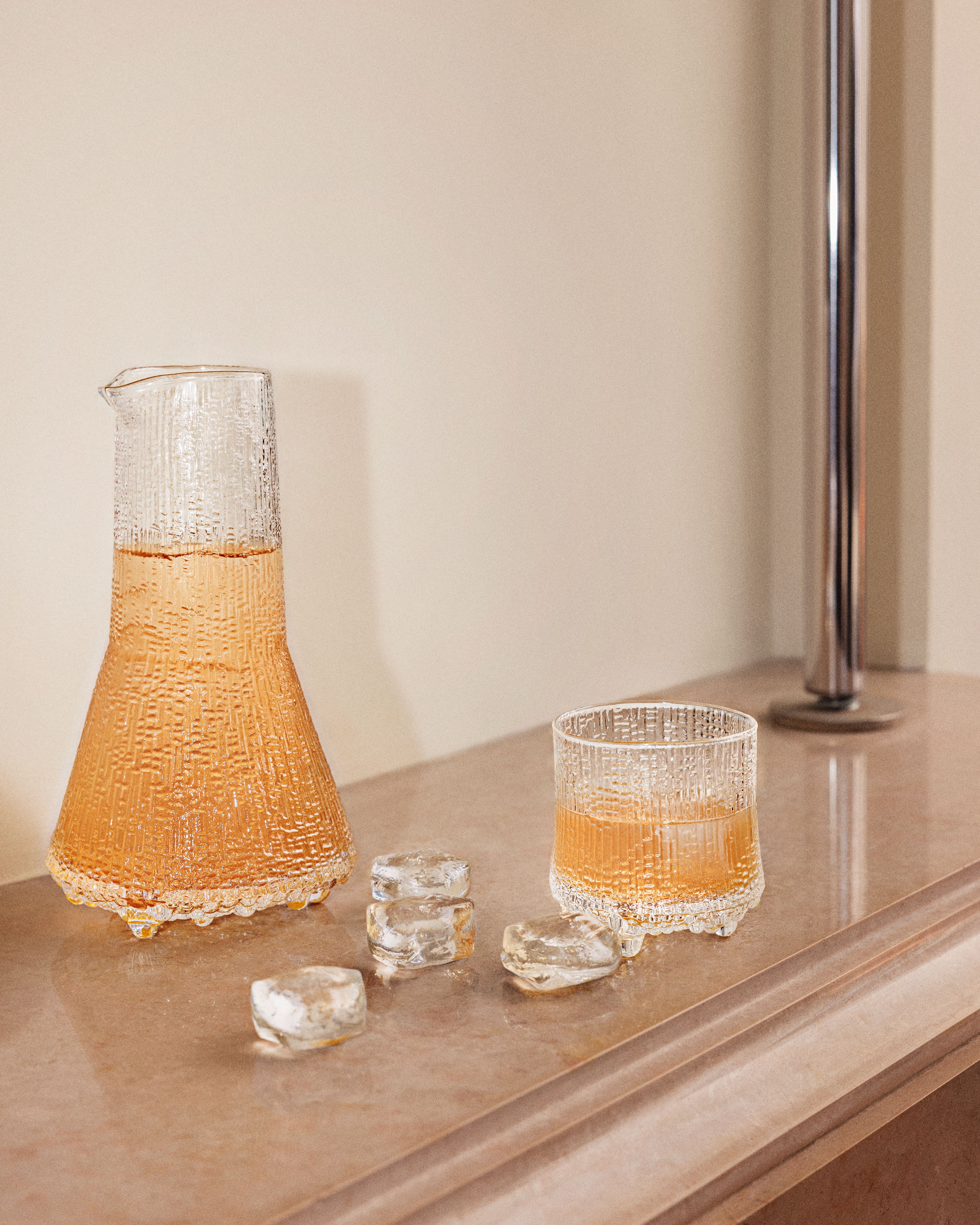 Teksturoitu lasinen karahvi ja siihen sopiva muki, jossa on meripihkanväristä nestettä, sulavien jääpalojen ympäröimänä vaalealla työtasolla.