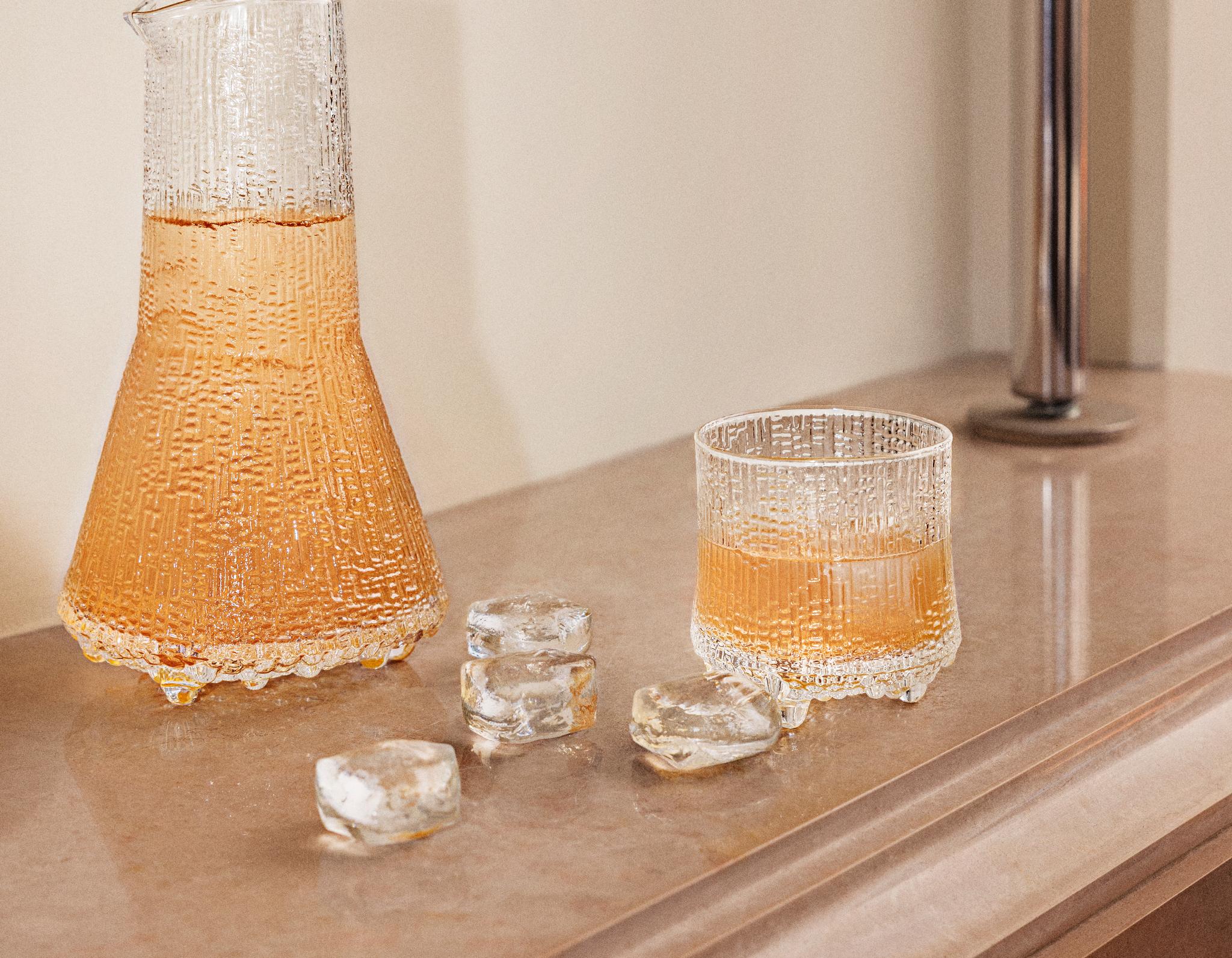 Teksturoitu lasinen karahvi ja siihen sopiva muki, jossa on meripihkanväristä nestettä, sulavien jääpalojen ympäröimänä vaalealla työtasolla.