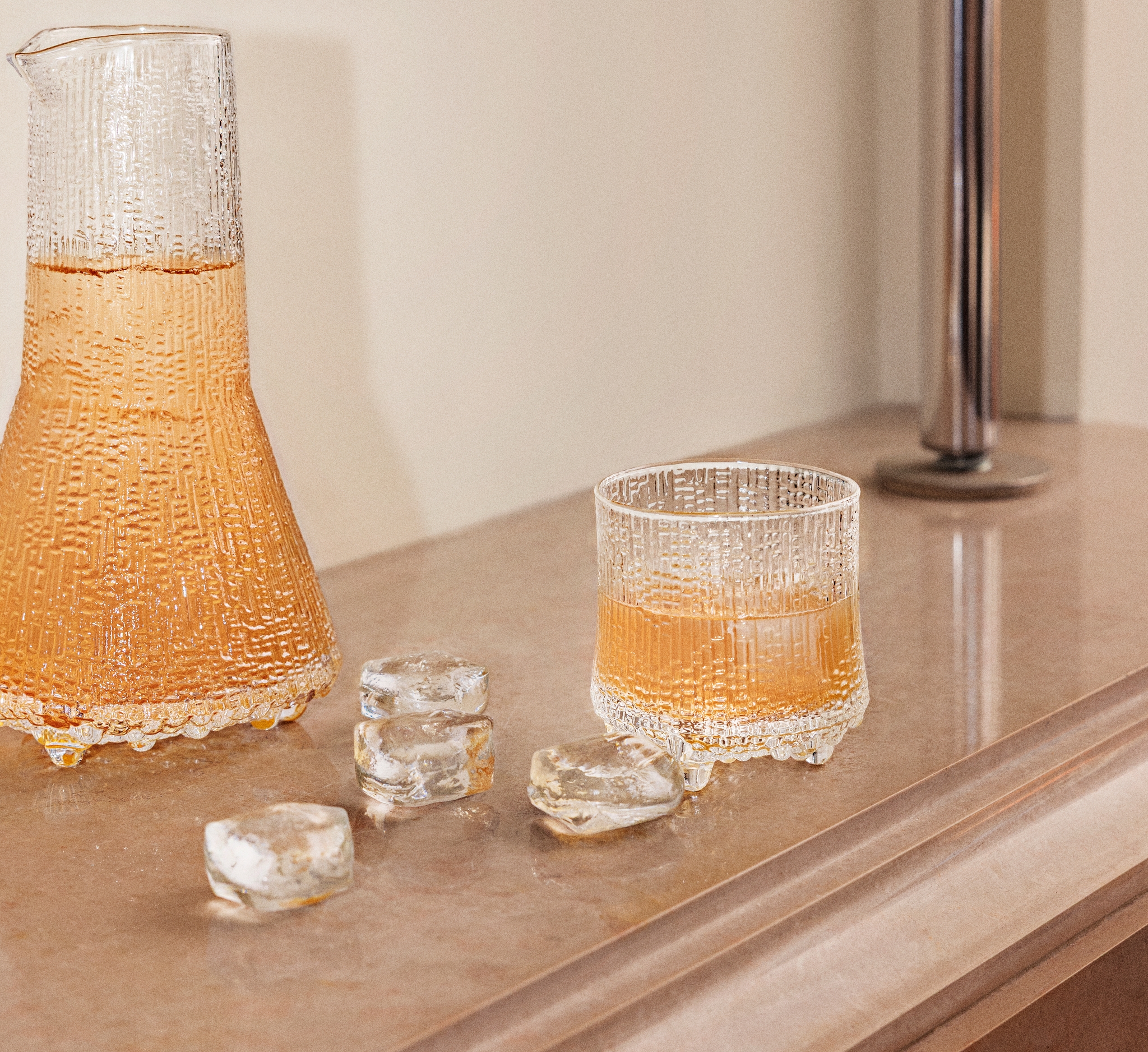 Teksturoitu lasinen karahvi ja siihen sopiva muki, jossa on meripihkanväristä nestettä, sulavien jääpalojen ympäröimänä vaalealla työtasolla.