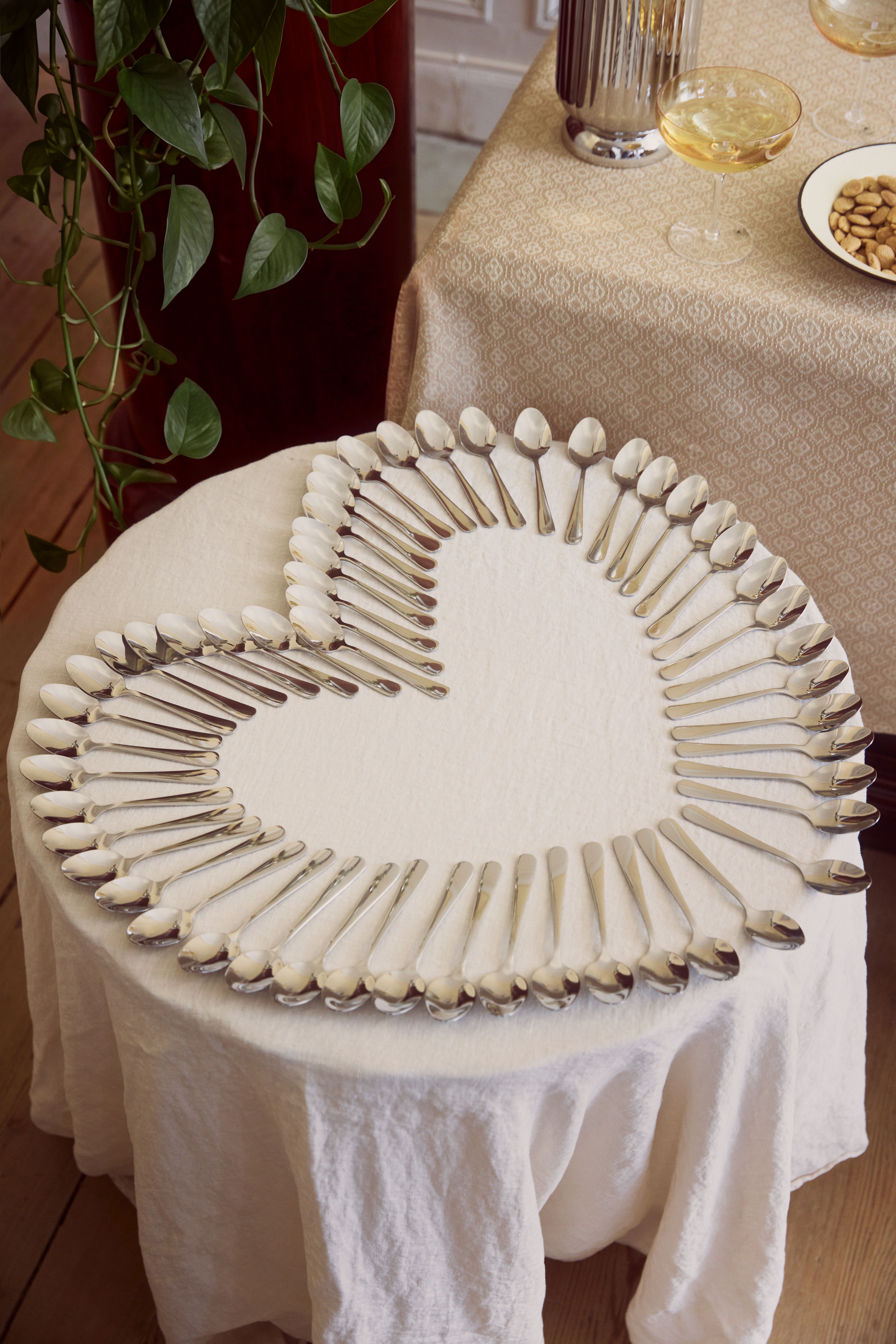 Silverfärgade skedar ligger ordnade i en hjärtform på ett runt, vitdukat bord vid en elegant dukning.