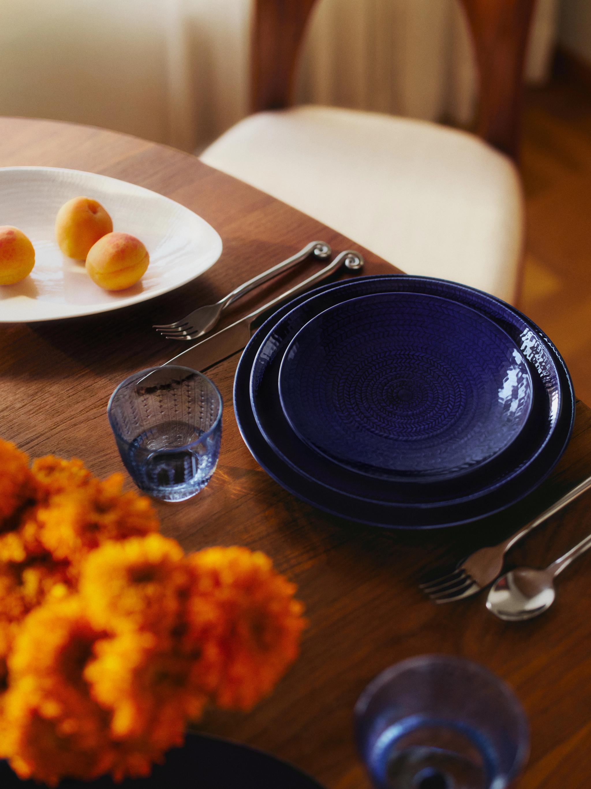 Ett matbord dukat med blå tallrikar, bestick, ett fat med aprikoser och orange blommor.
