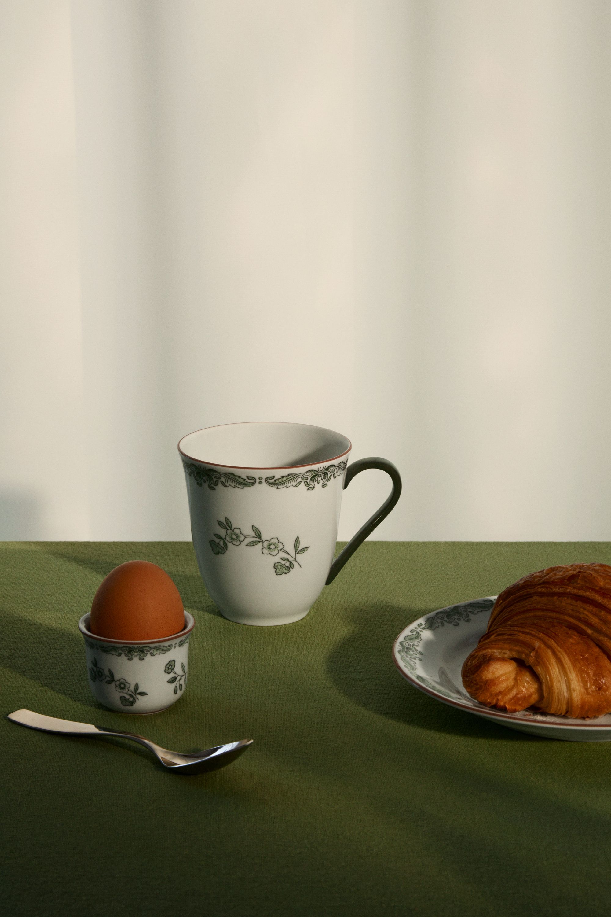 Frukostdukning med blommig porslinskopp, kokt ägg och croissant på en grön duk.