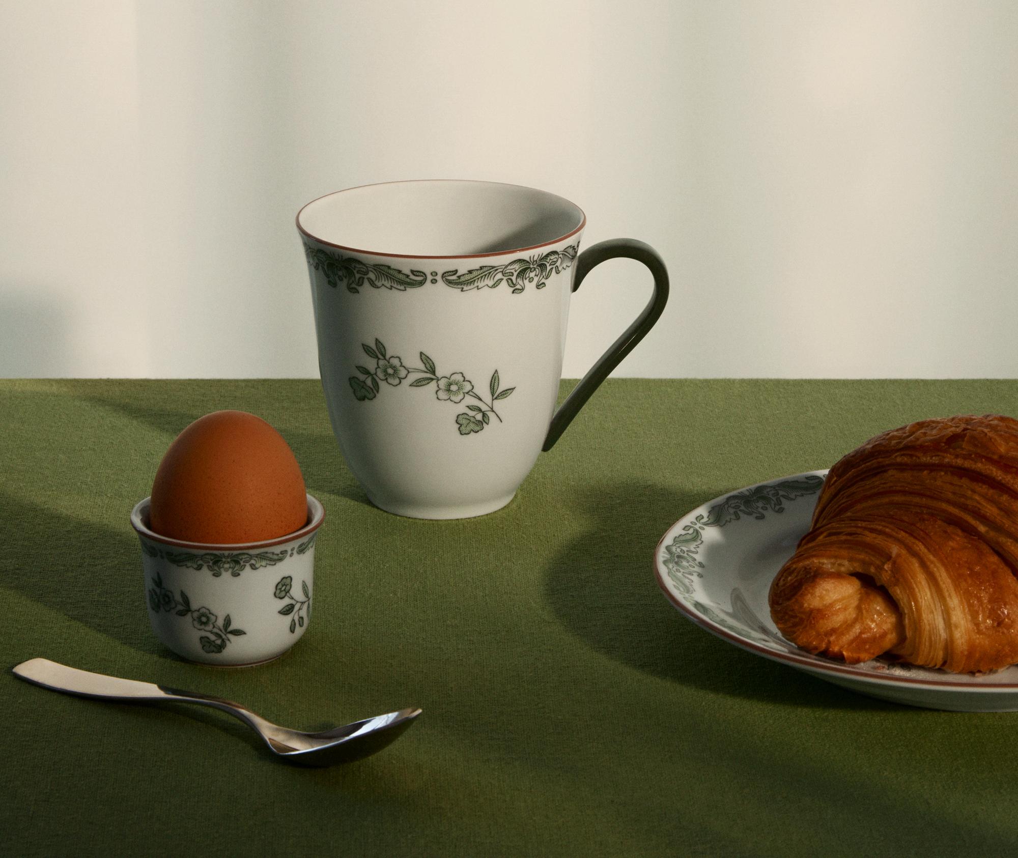 Frokost med croissant, kokt egg og kaffekopp på en grønn duk.