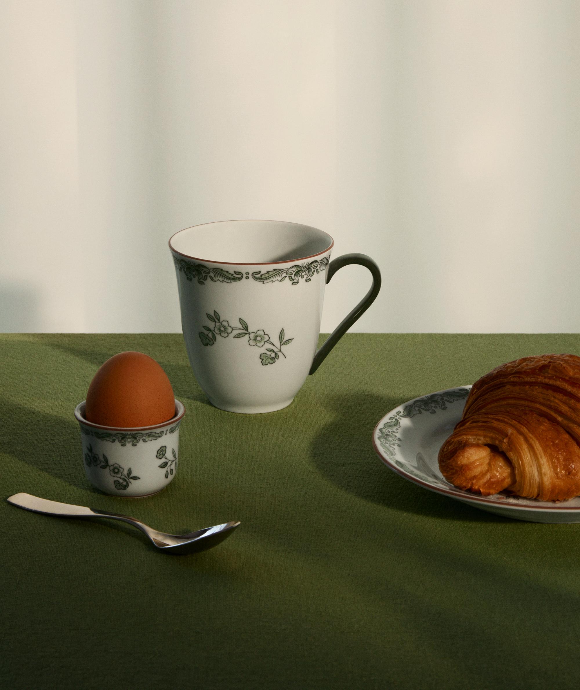 Frokostbord med kaffekopp, kokt egg og croissant på grønn duk.