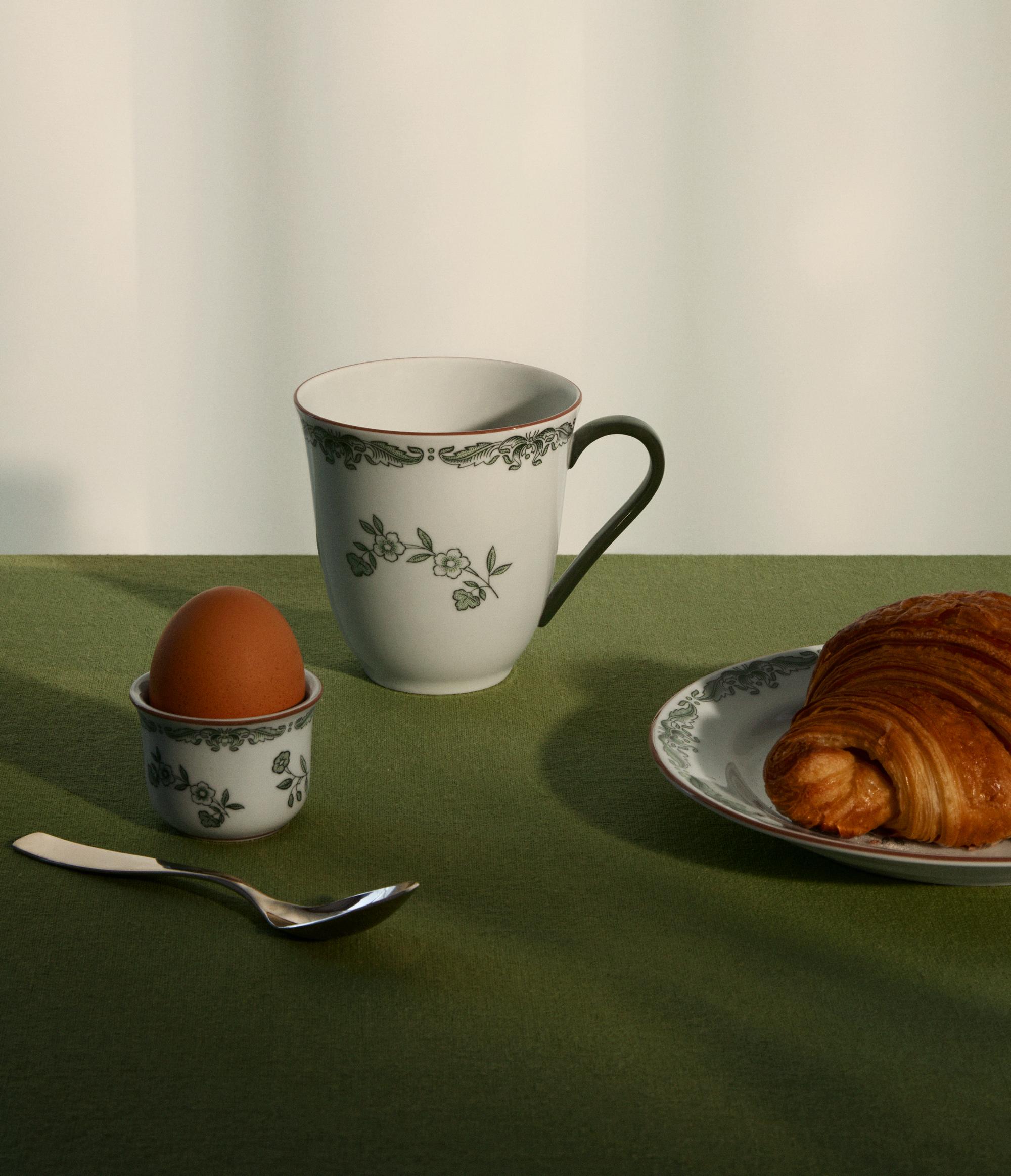 Stilren frukostdukning med kaffekopp, kokt ägg och croissant på grön duk.