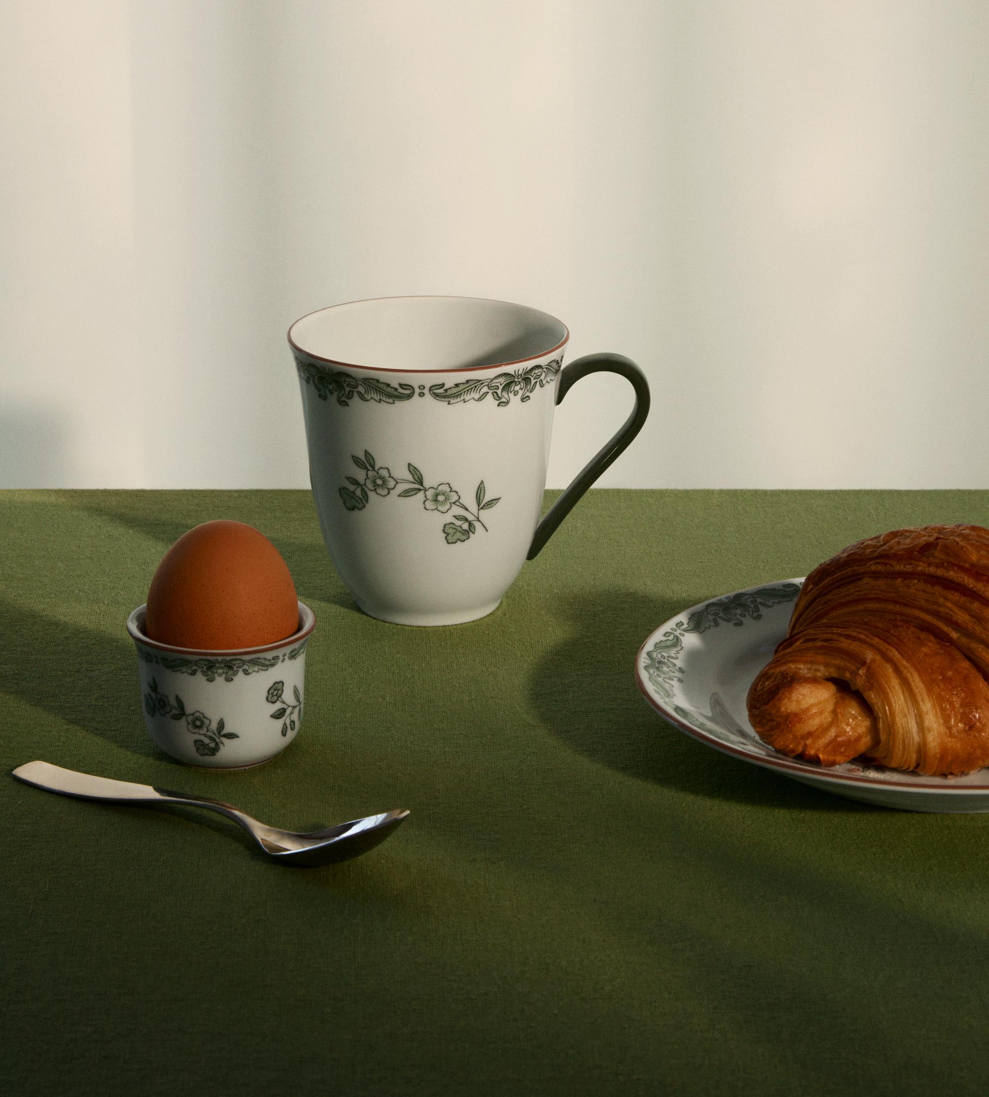 Frukostdukning med croissant, kokt ägg och blommönstrad kaffekopp på en grön duk.