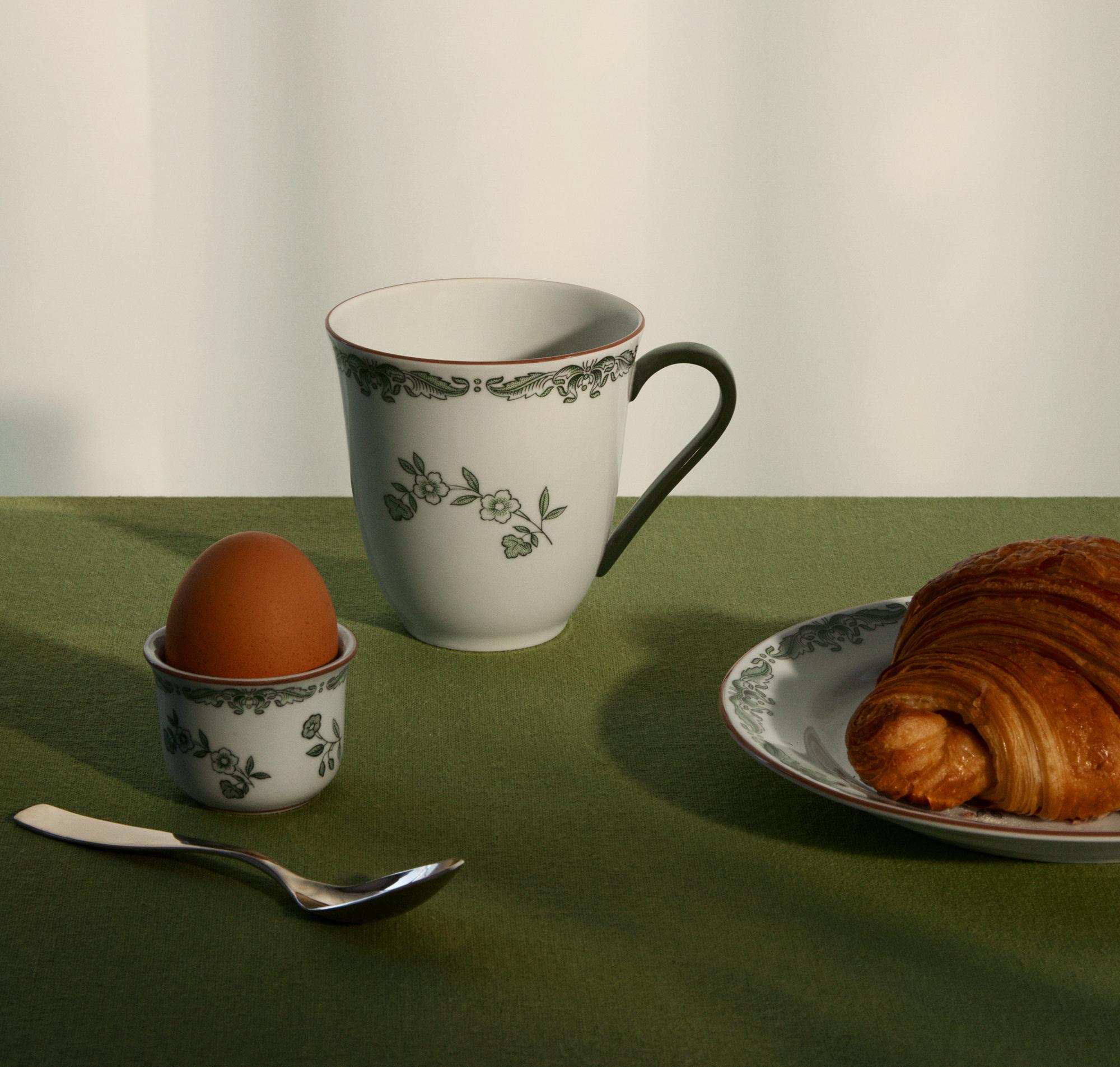 Frukostdukning med blommig porslinskopp, kokt ägg och croissant på en grön duk.