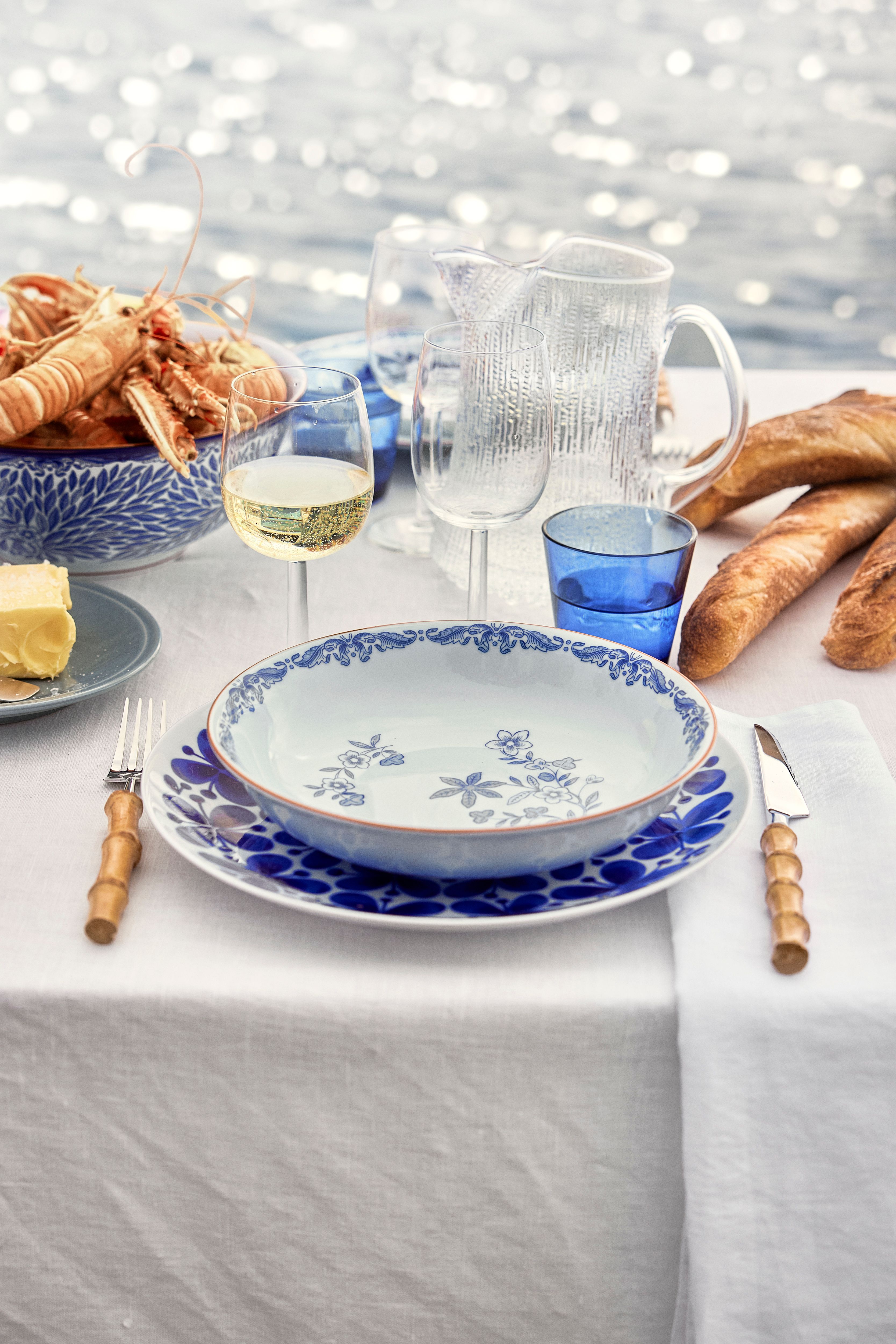 Somrig dukning vid havet med blåvitt porslin, skaldjur, baguetter och vitt vin på vit linneduk.