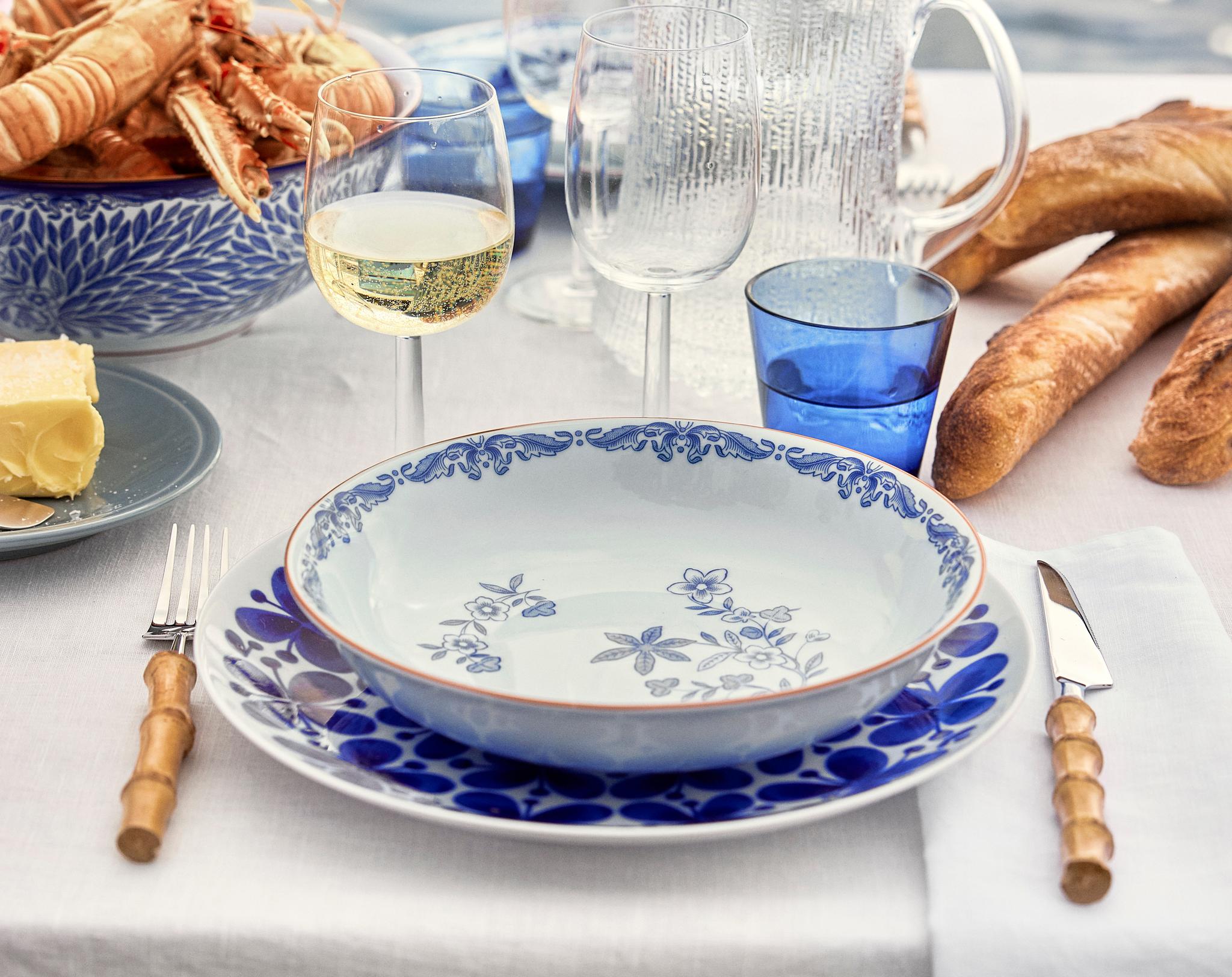 Somrig dukning vid havet med blåvitt porslin, skaldjur, baguetter och vitt vin på vit linneduk.