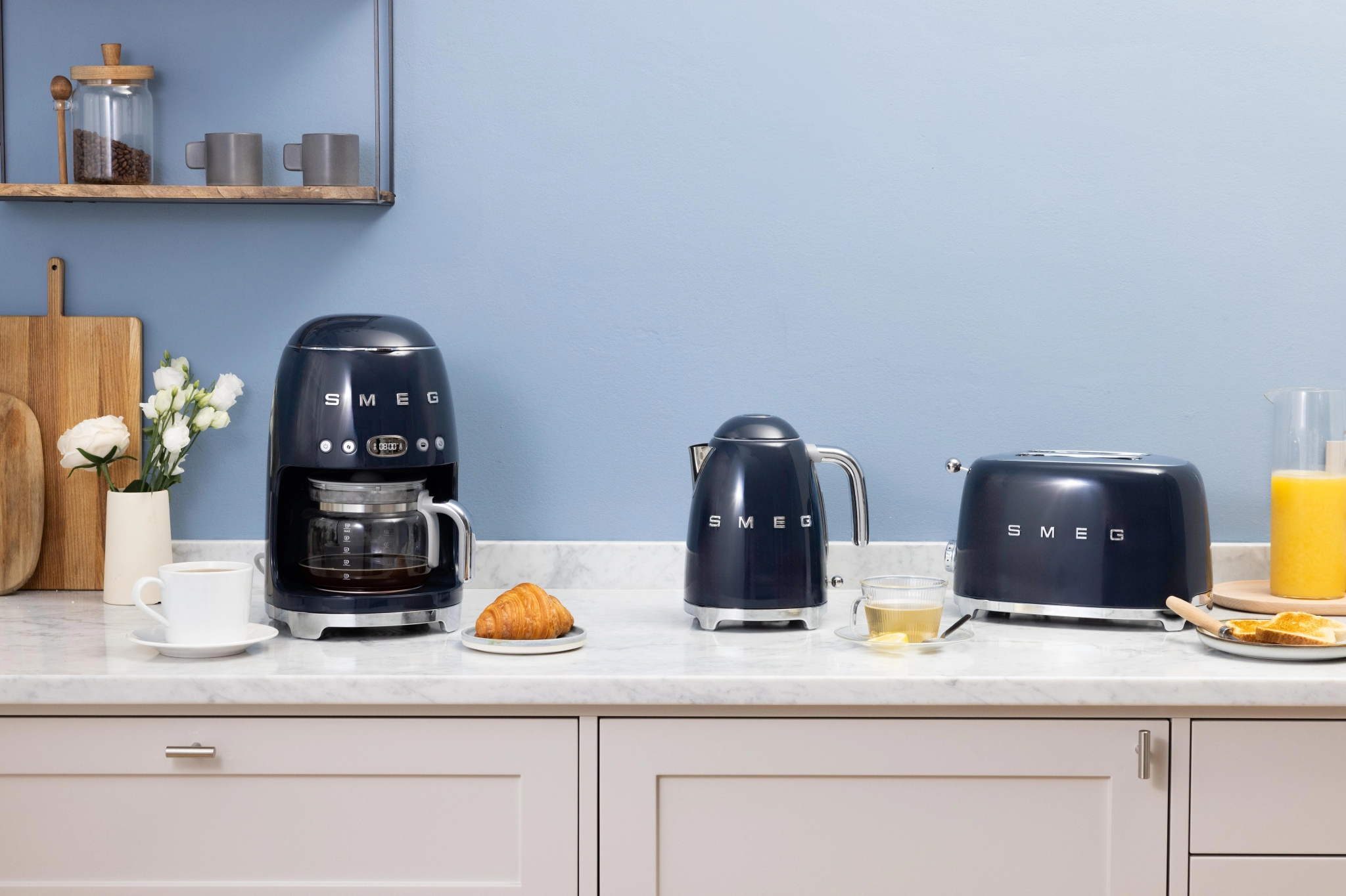 Marineblå Smeg kaffetrakter, vannkoker og brødrister på en kjøkkenbenk med frokostvarer.