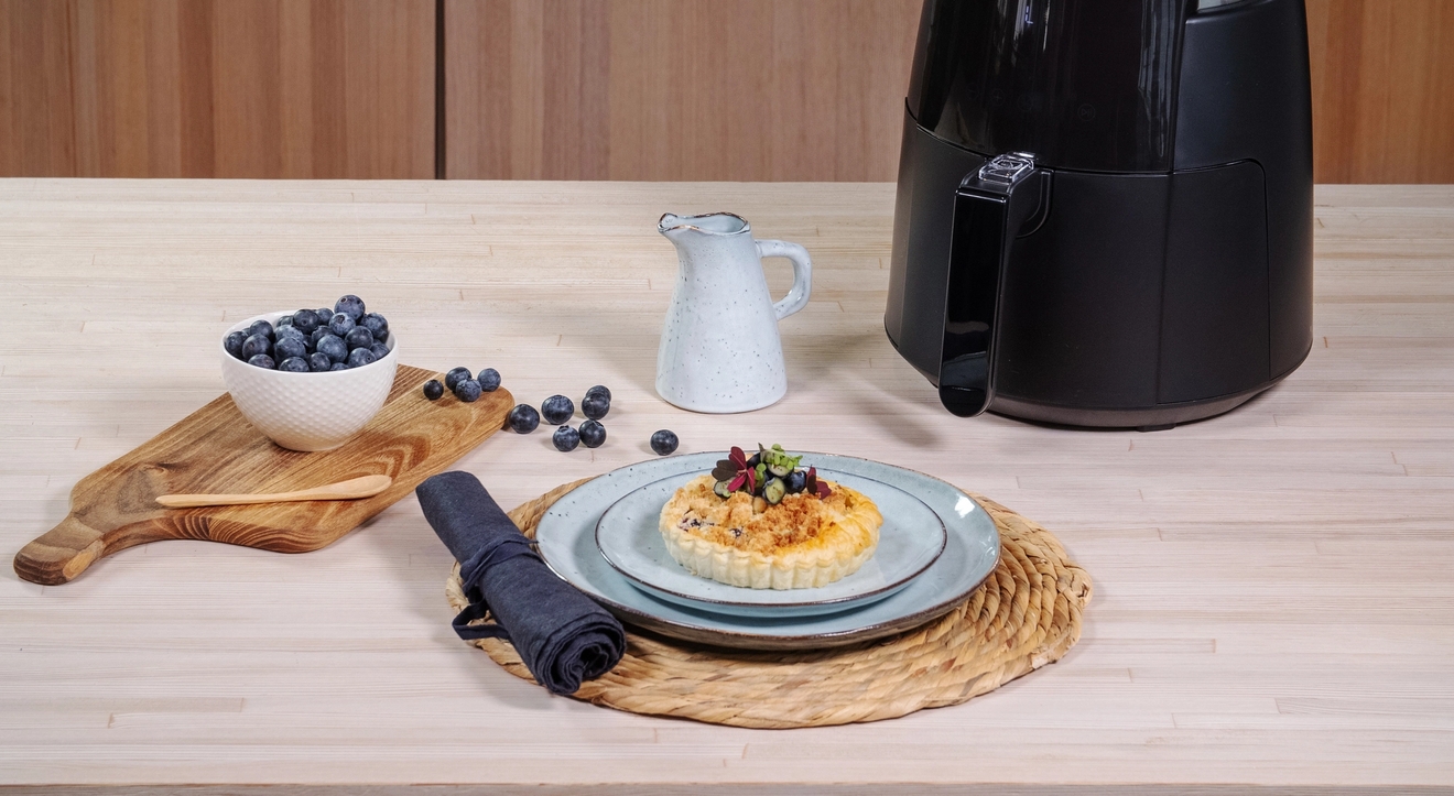 Pieni airfryer keittiön työtasolla ja makea piirakka vaaleansinisellä lautasella mustikoiden kera