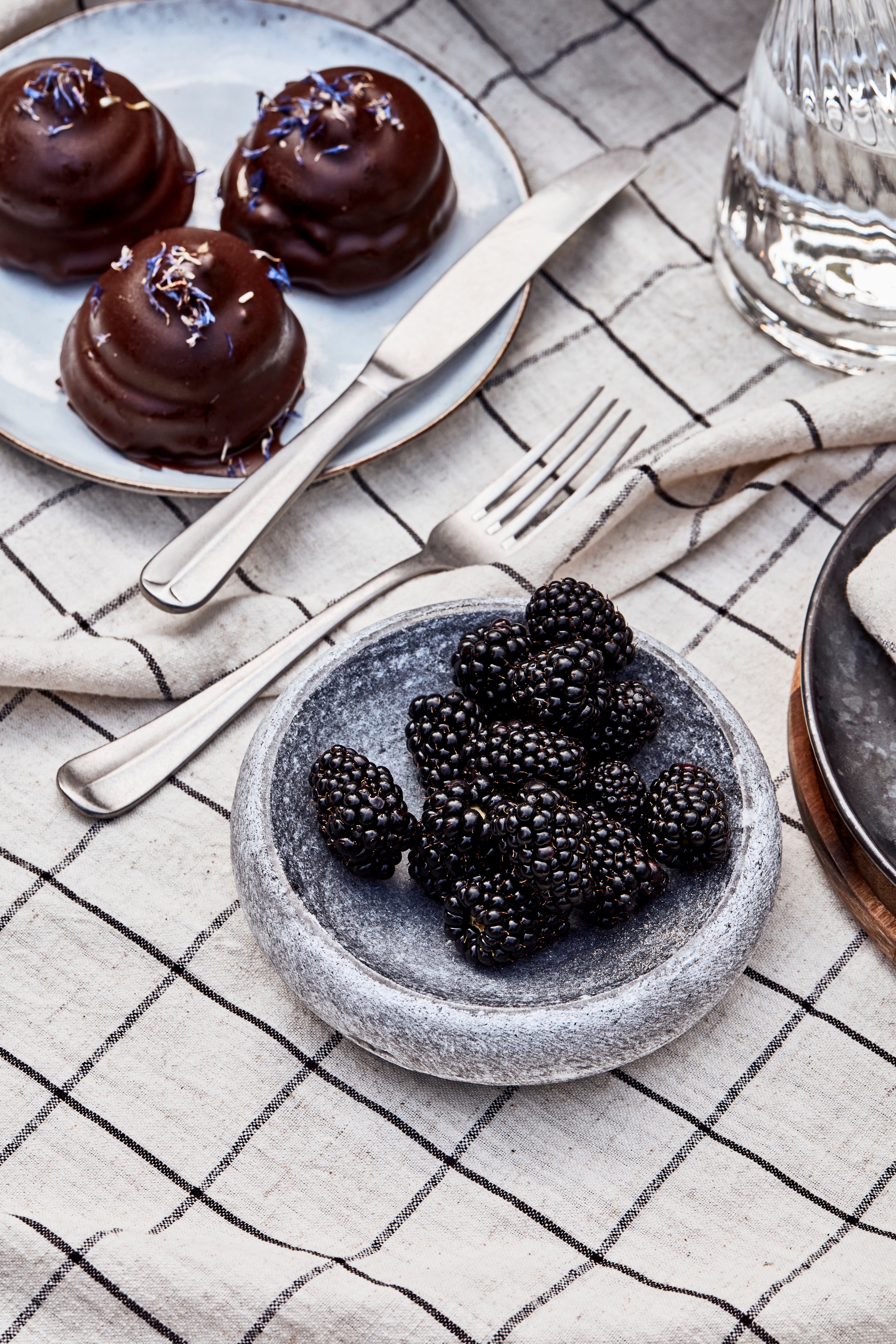 Tre chokladbakelser med blomströssel, en skål med björnbär och bestick på en rutig duk