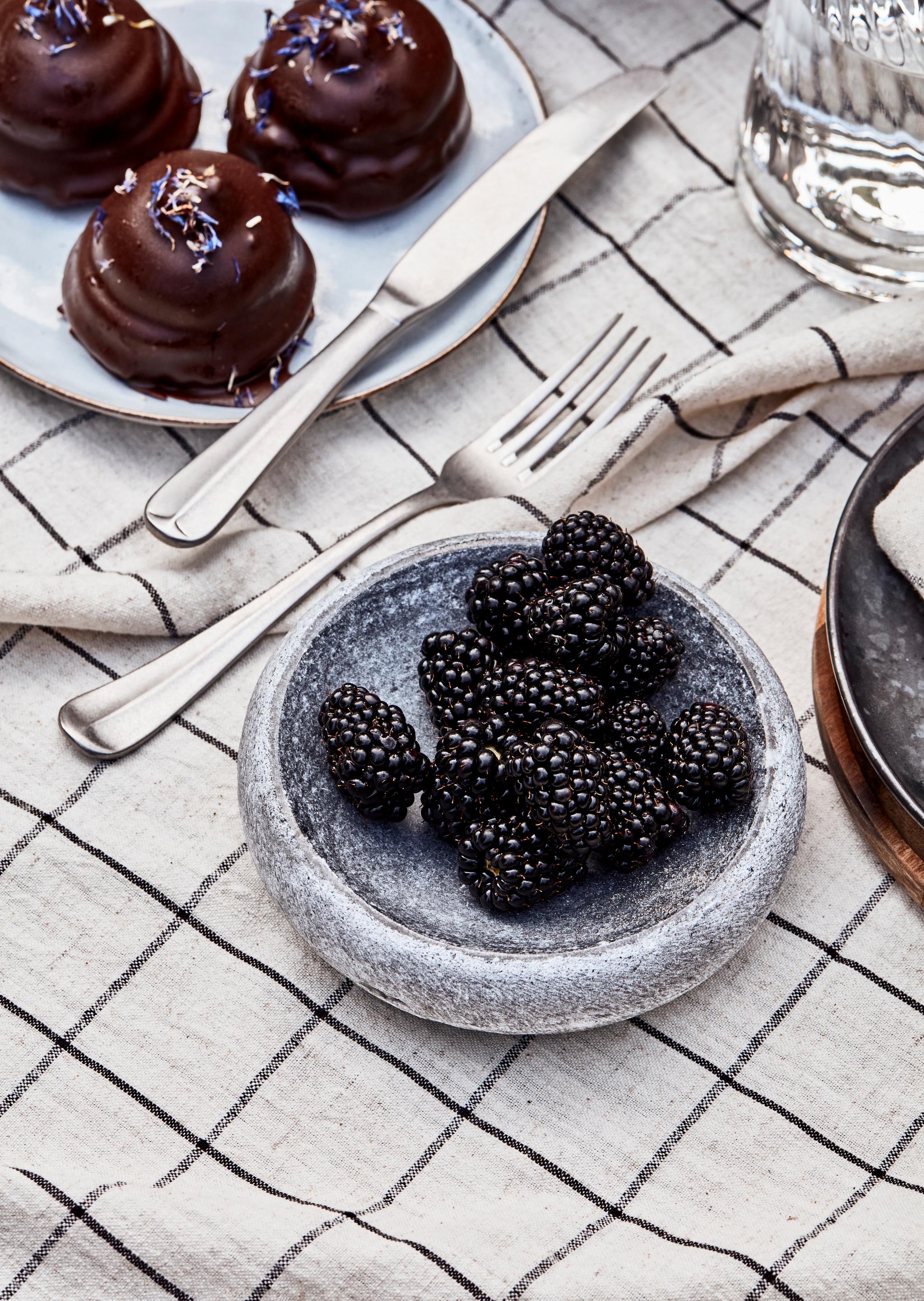 Tre chokladbakelser med blomströssel, en skål med björnbär och bestick på en rutig duk