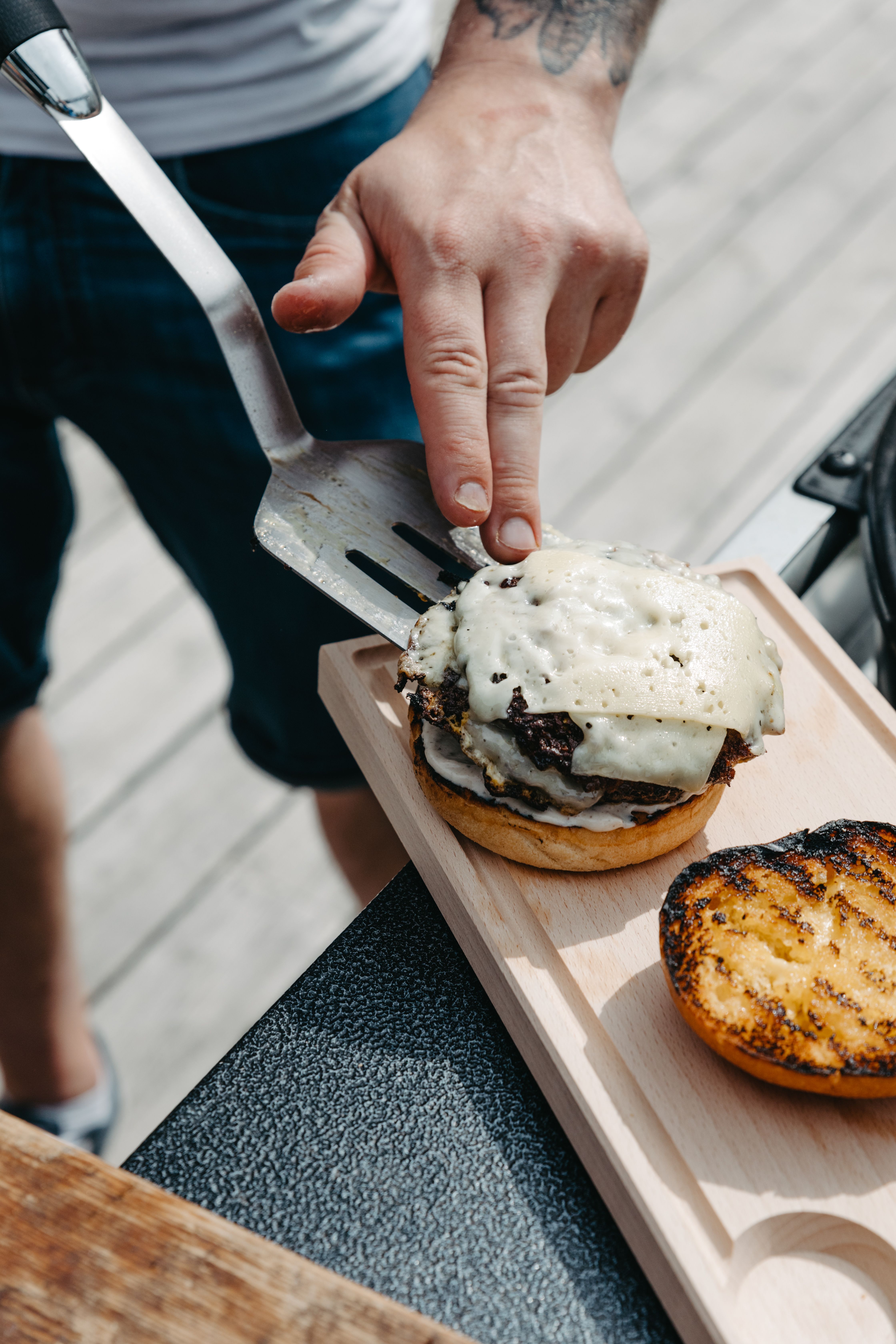 En person monterar en grillad hamburgare med smält ost på en brödskiva utomhus vid grillen.