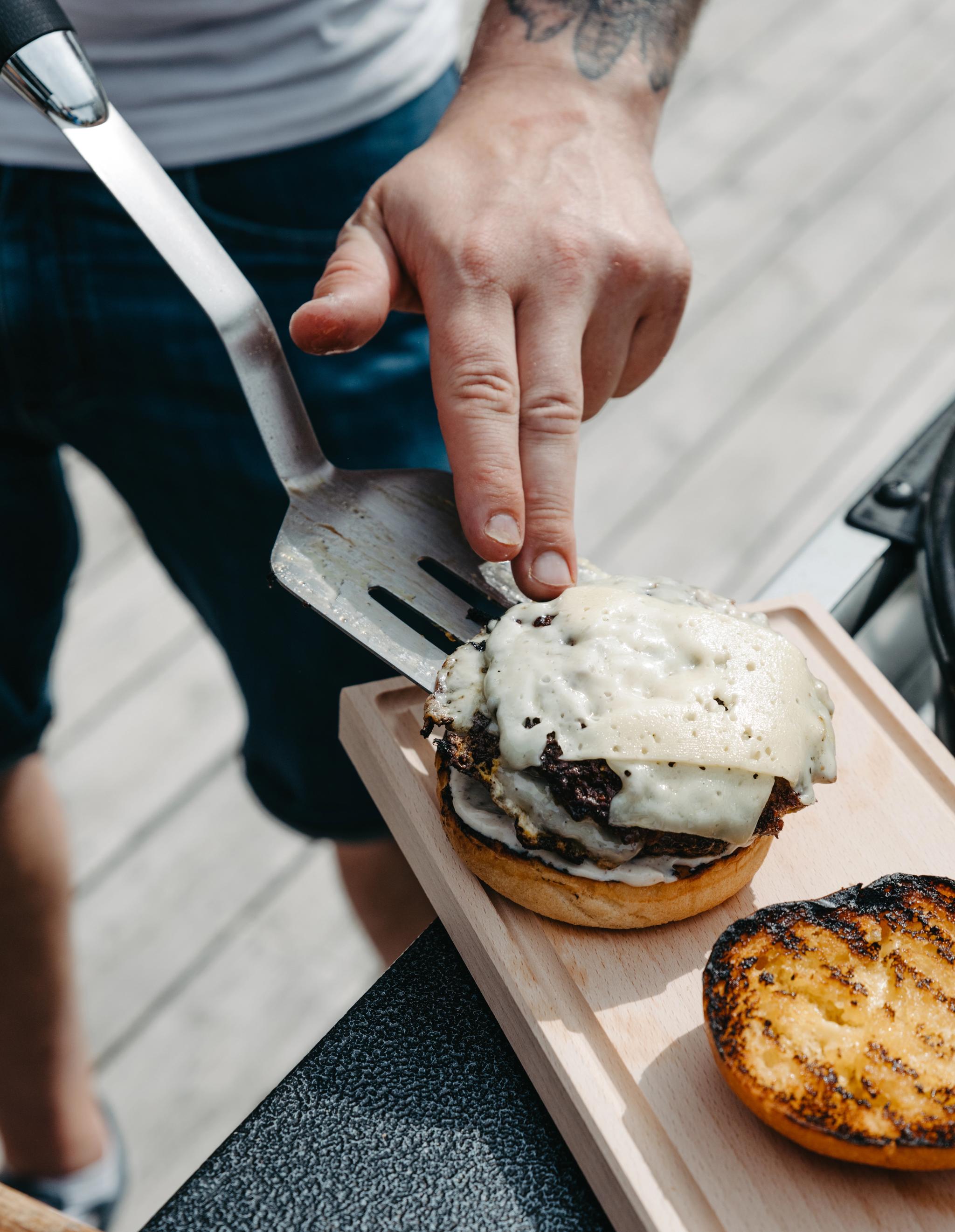 En person monterar en grillad hamburgare med smält ost på en brödskiva utomhus vid grillen.