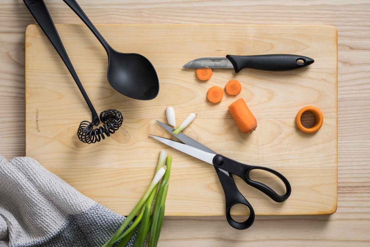 Fiskars keittiövälineet kuten vispilä kauha sakset ja kuorimaveitsi puisella leikkuulaudalla vihannesten kera