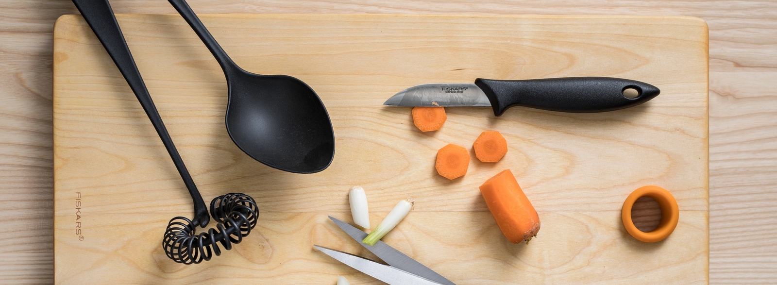 Fiskars keittiövälineet kuten vispilä kauha sakset ja kuorimaveitsi puisella leikkuulaudalla vihannesten kera