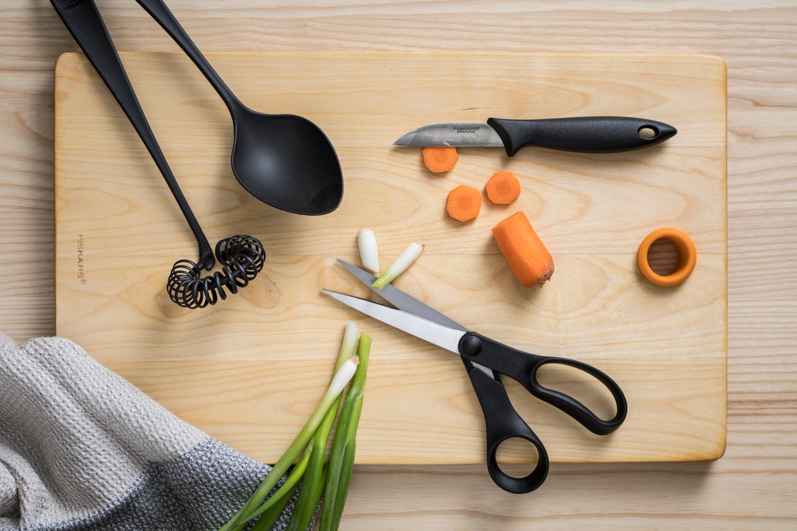 Fiskars keittiövälineet kuten vispilä kauha sakset ja kuorimaveitsi puisella leikkuulaudalla vihannesten kera