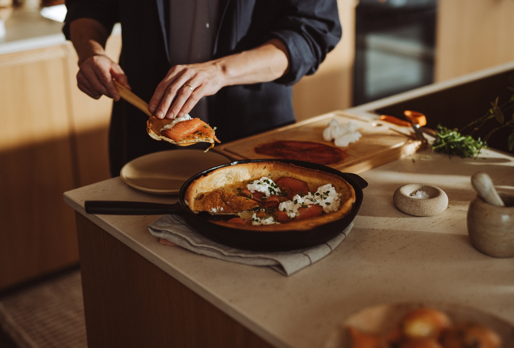 En person serverar en ugnsbakad pannkaka med lax, färskost och örter i ett varmt kök.