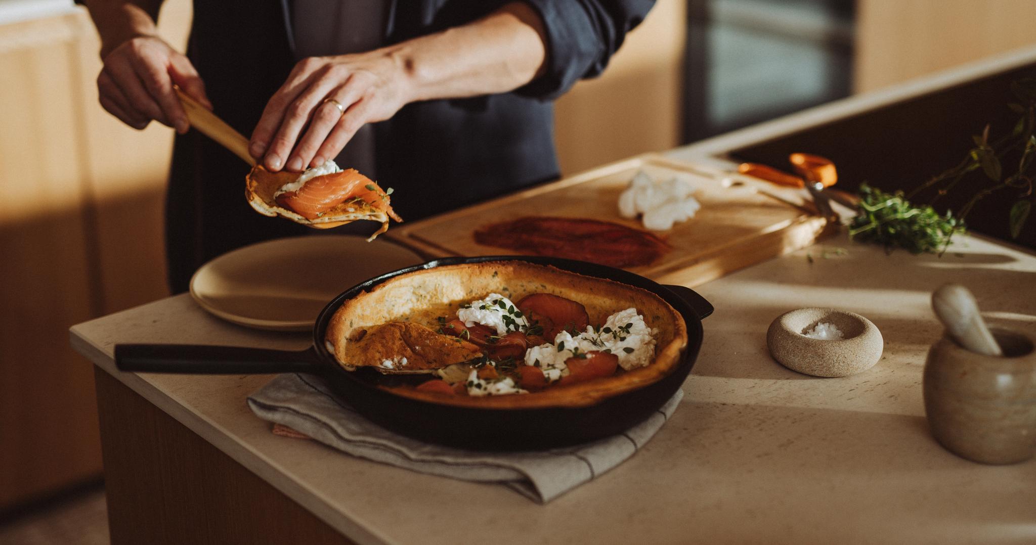 En person garnerar en holländsk pannkaka med lax och ost i ett kök.