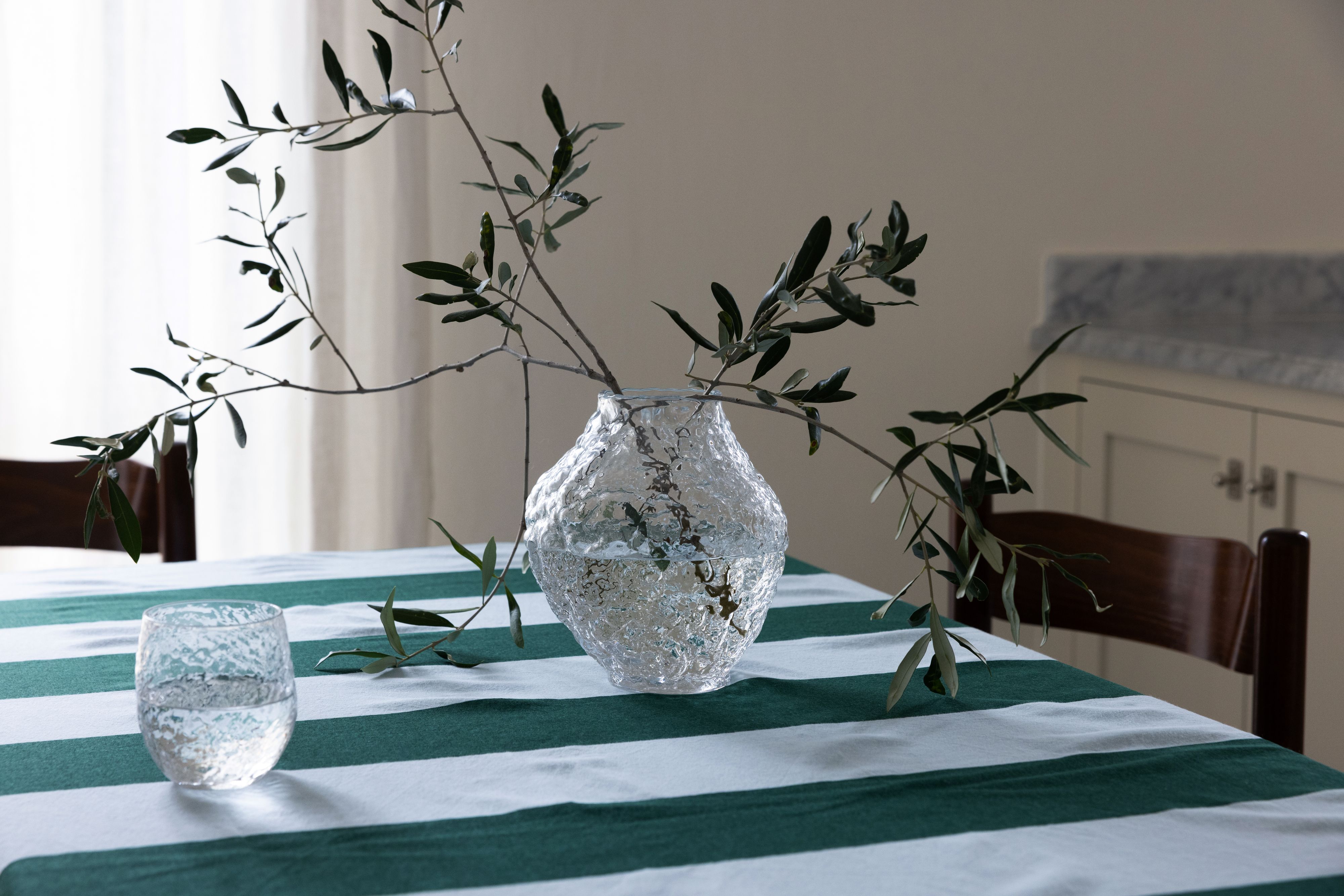 Textured glass vase with olive branches and a matching glass on a green and white striped tablecloth.