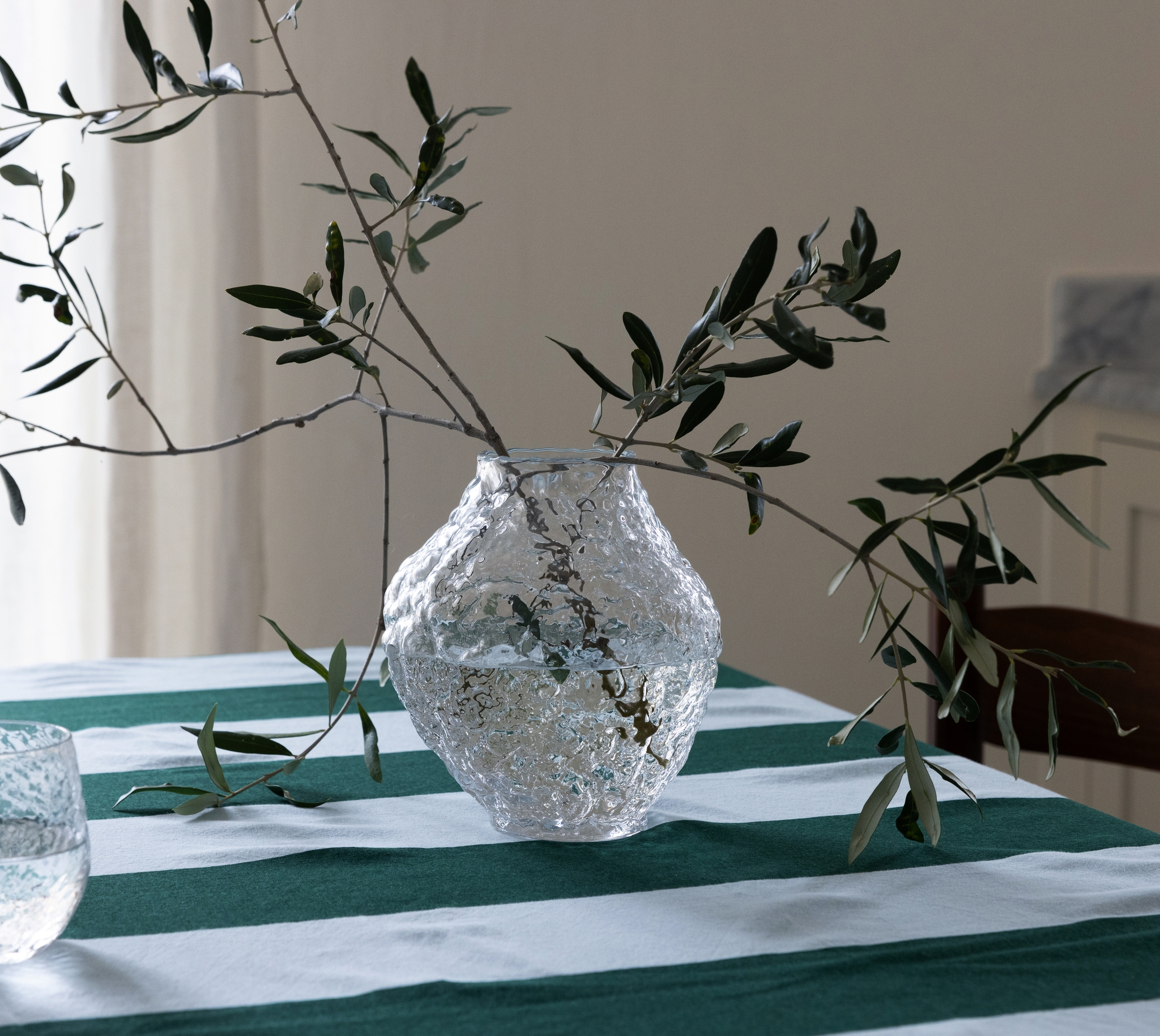 Textured glass vase with olive branches on a green and white striped tablecloth, with a matching tumbler.