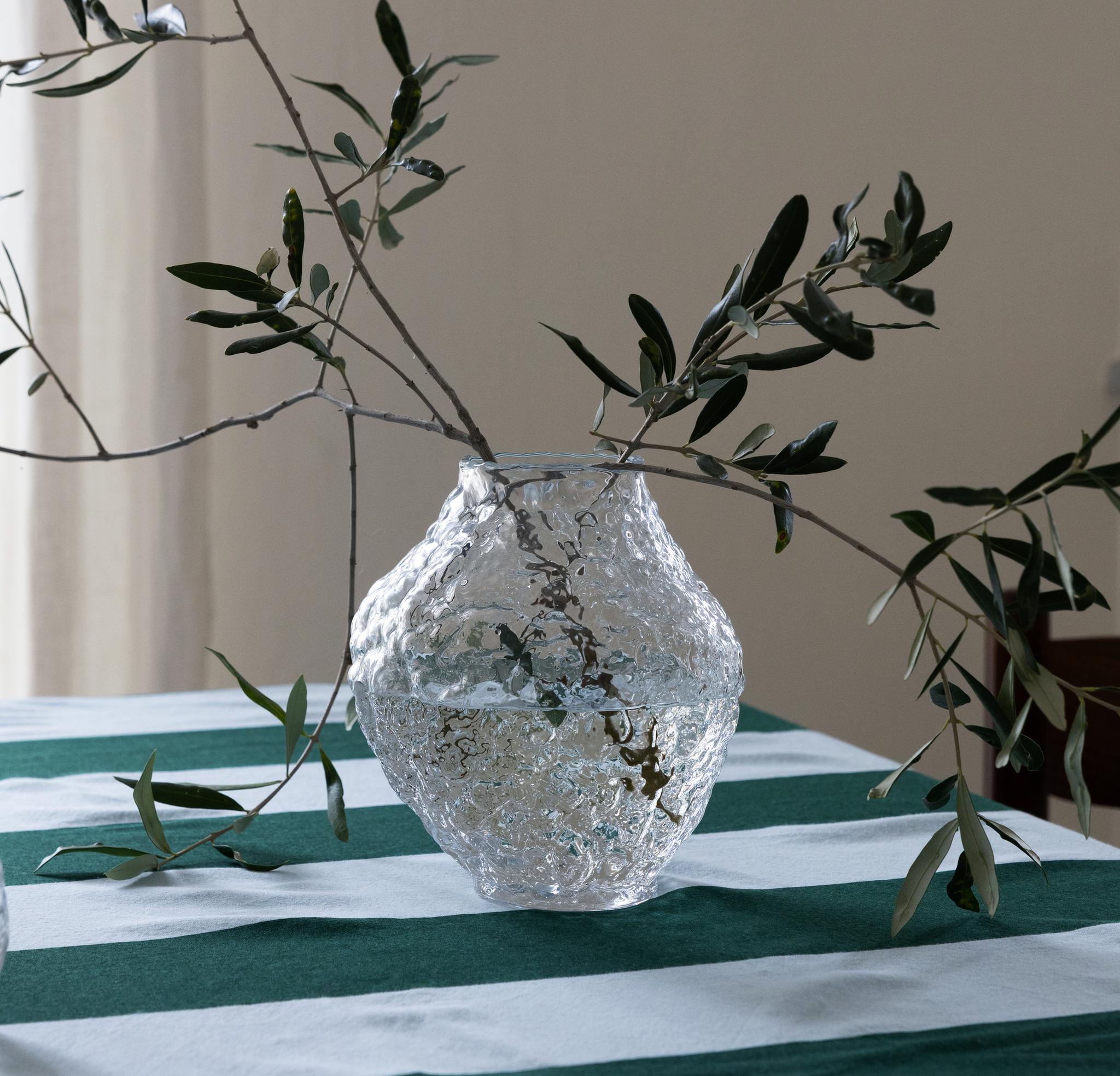 Textured glass vase with olive branches and a matching glass on a green and white striped tablecloth.