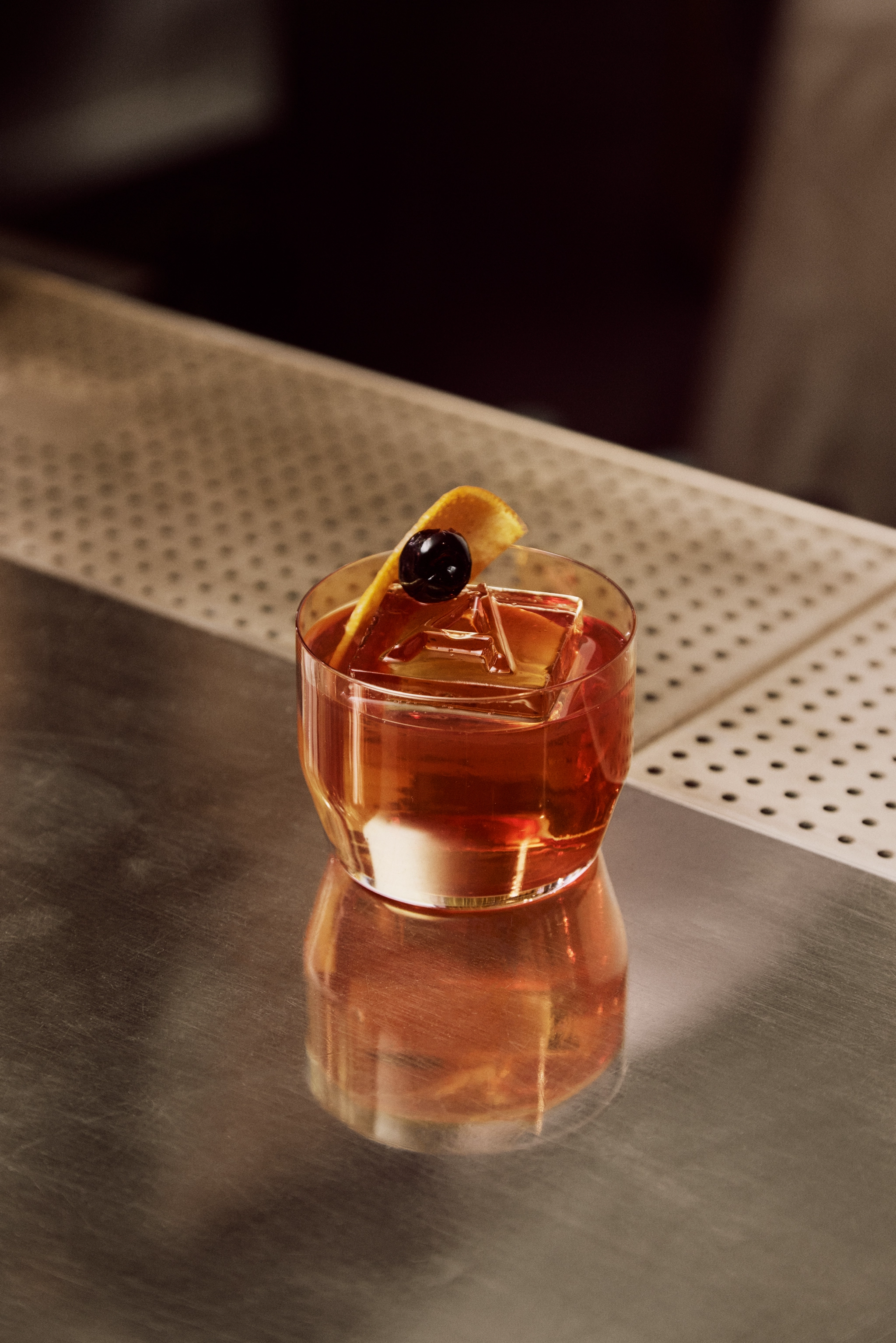 Bärnstensfärgad cocktail med is, apelsinskal och körsbär på en blank bardisk.