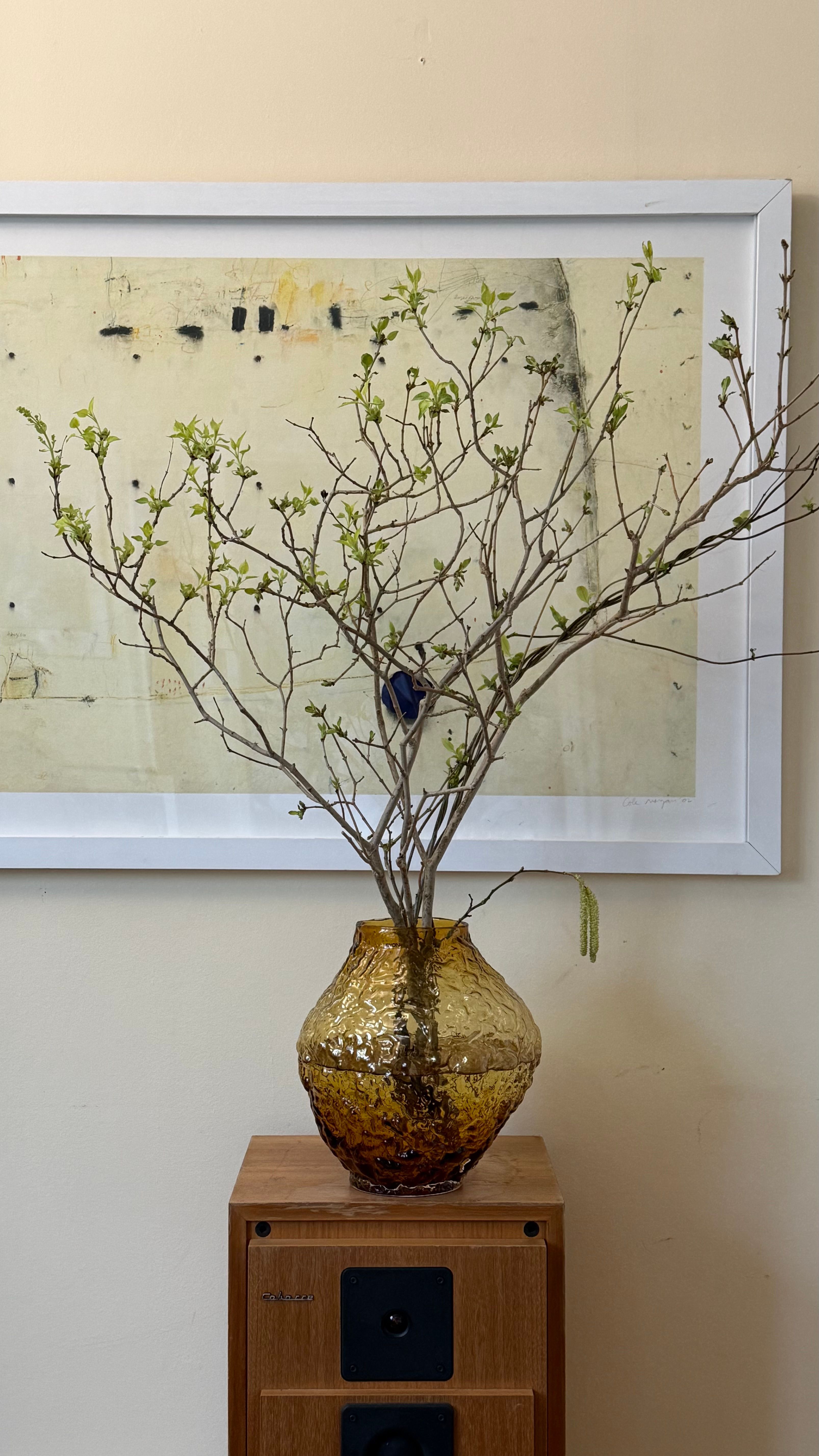 Spring branches with new green leaves in a textured amber vase on a wooden speaker, in front of a framed abstract artwork.