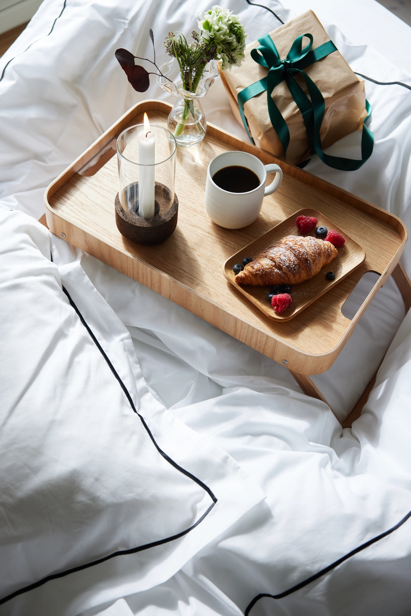 En frukostbricka i trä med en croissant, kaffe, ett tänt ljus, blommor och en inslagen present på vita sängkläder.
