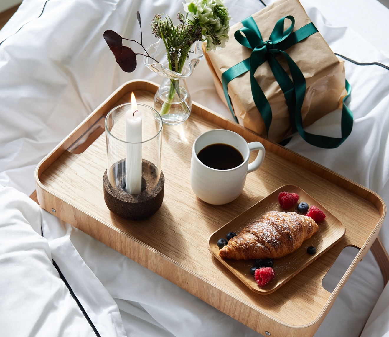 Breakfast in bed with coffee, a croissant, berries, a lit candle, and flowers on a wooden tray, next to a gift on white bedding.