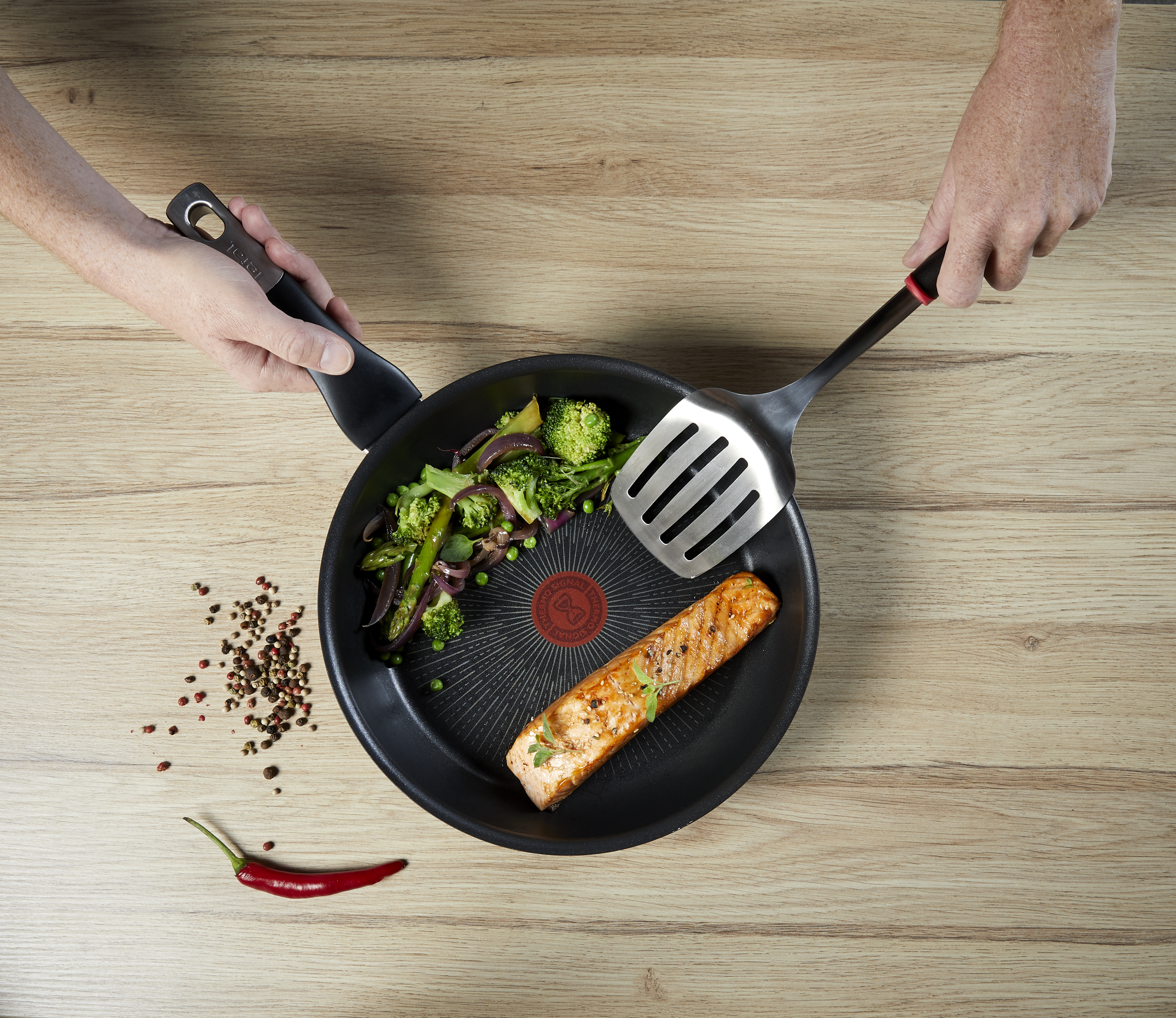 Kypsennettyä lohta ja sekavihanneksia T-paistinpannulla, kädet pitelevät pannua ja lastaa puupöydällä.