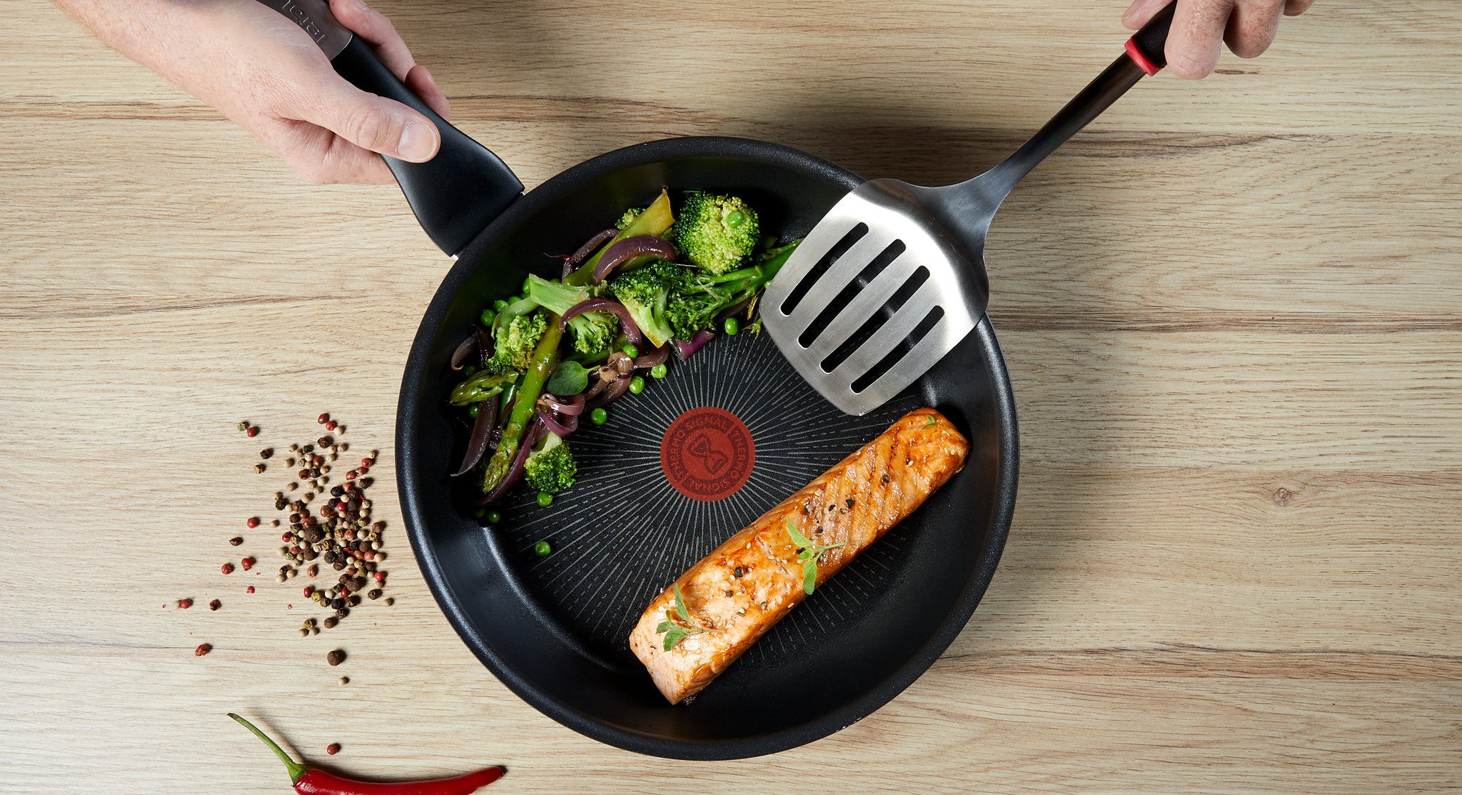 Kypsennettyä lohta ja sekavihanneksia T-paistinpannulla, kädet pitelevät pannua ja lastaa puupöydällä.