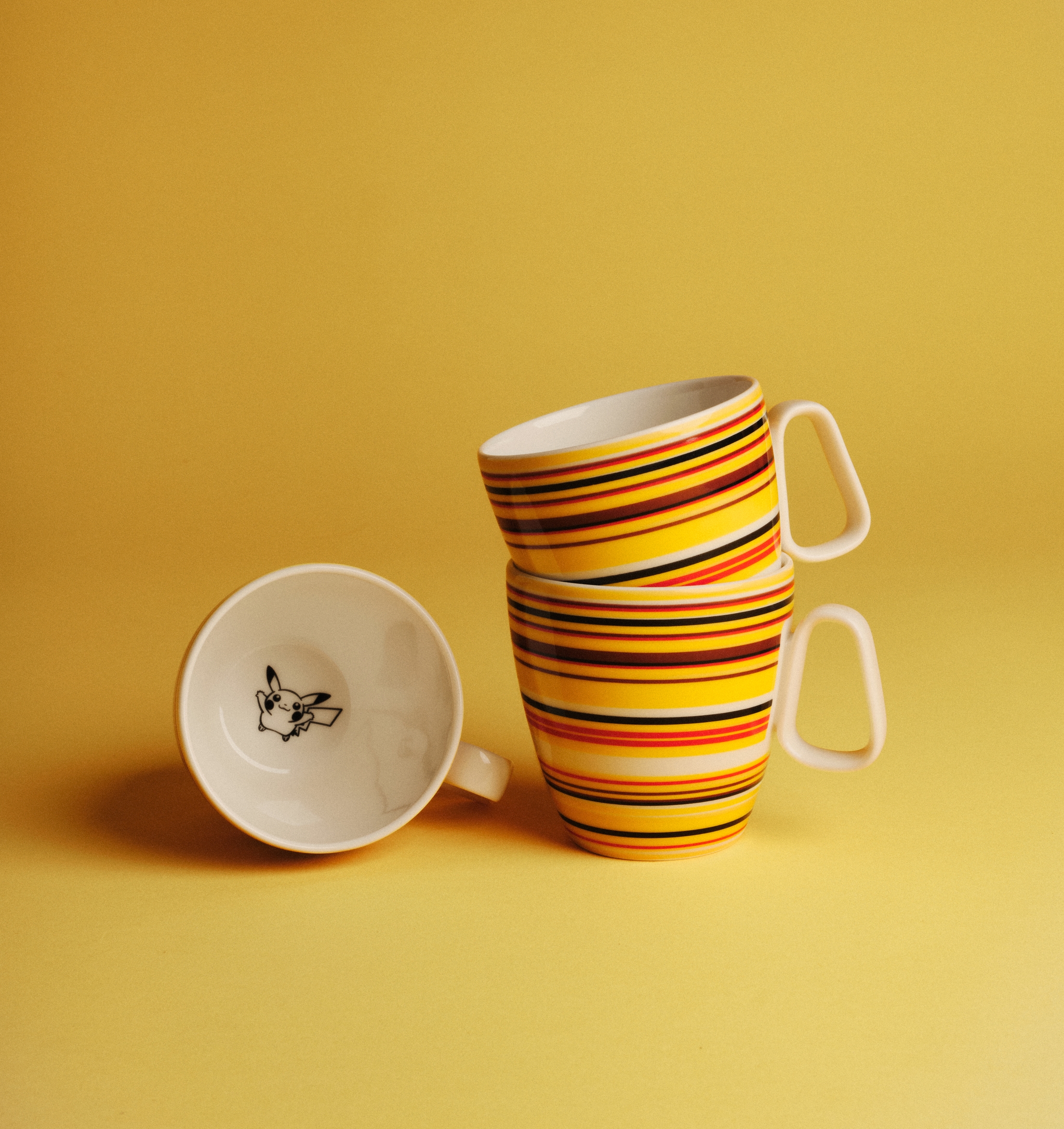 Tre färgglada randiga kaffekoppar, en liggande med Pikachu-motiv, mot en gul bakgrund.