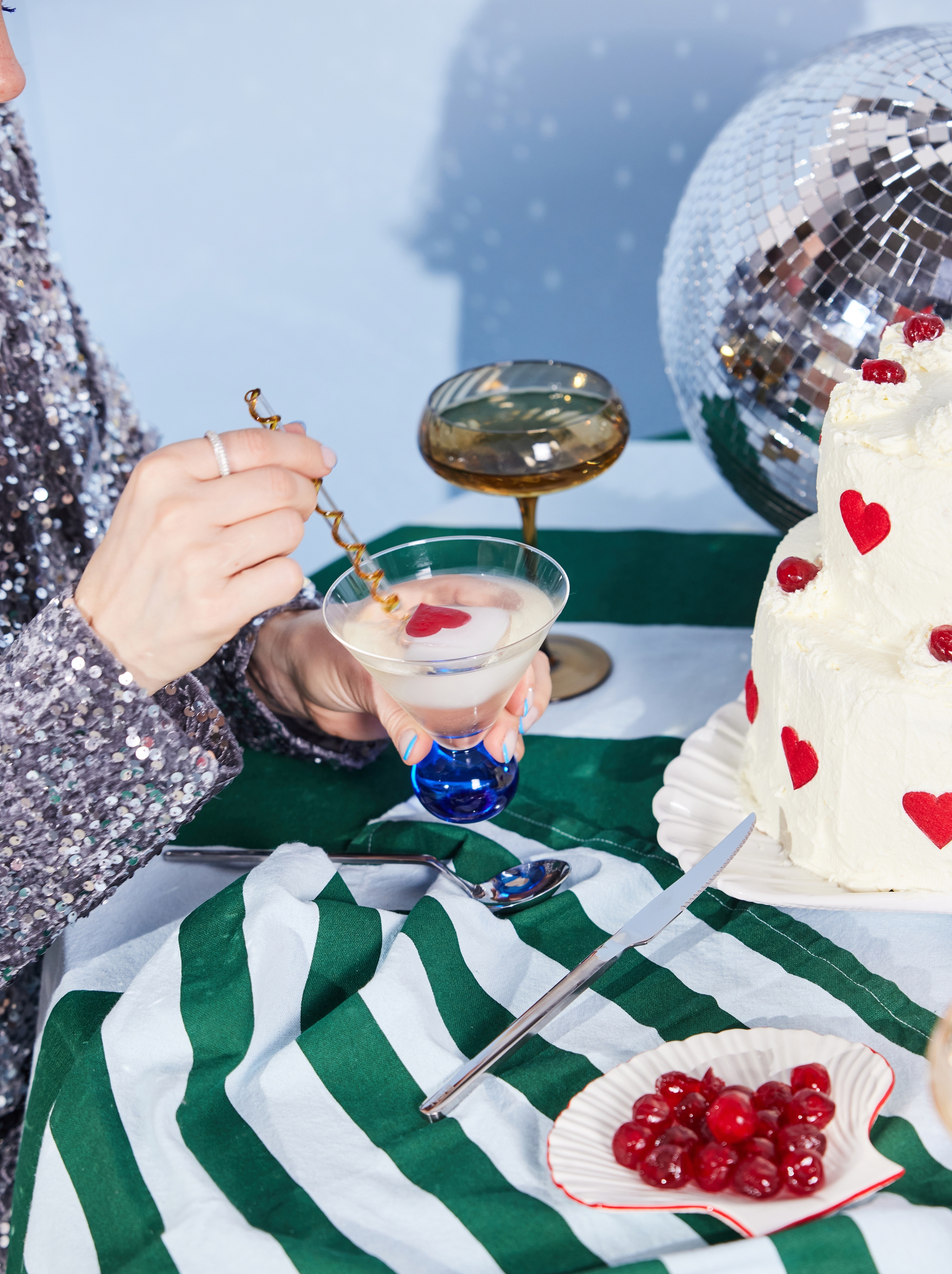 En person i en paljettopp rör om en cocktail med en hjärtformad garnering på ett bord med en vit tårta dekorerad med röda hjärtan, en skål med körsbär och en discokula