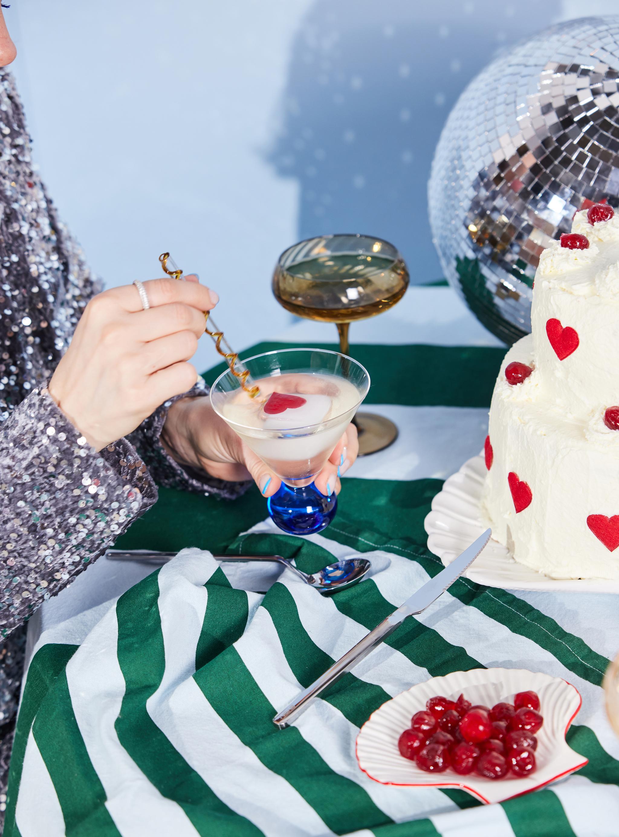 En person i en paljettopp rör om en cocktail med en hjärtformad garnering på ett bord med en vit tårta dekorerad med röda hjärtan, en skål med körsbär och en discokula