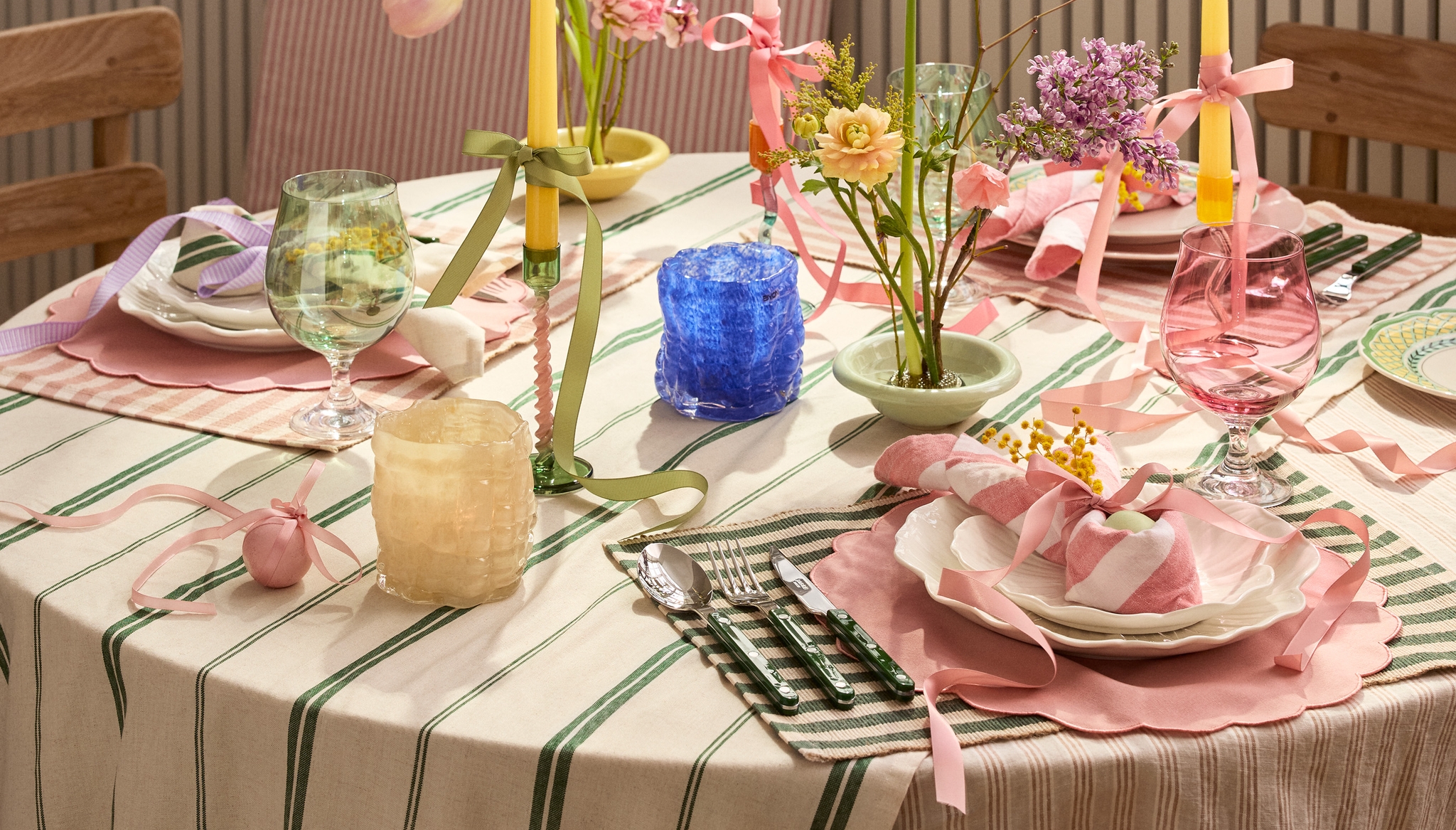 Kauniisti katettu kevätpöytä raidallisella pöytäliinalla, vaaleanpunaisilla tableteilla, kukka-asetelmilla ja pastellivärisillä kynttilöillä.
