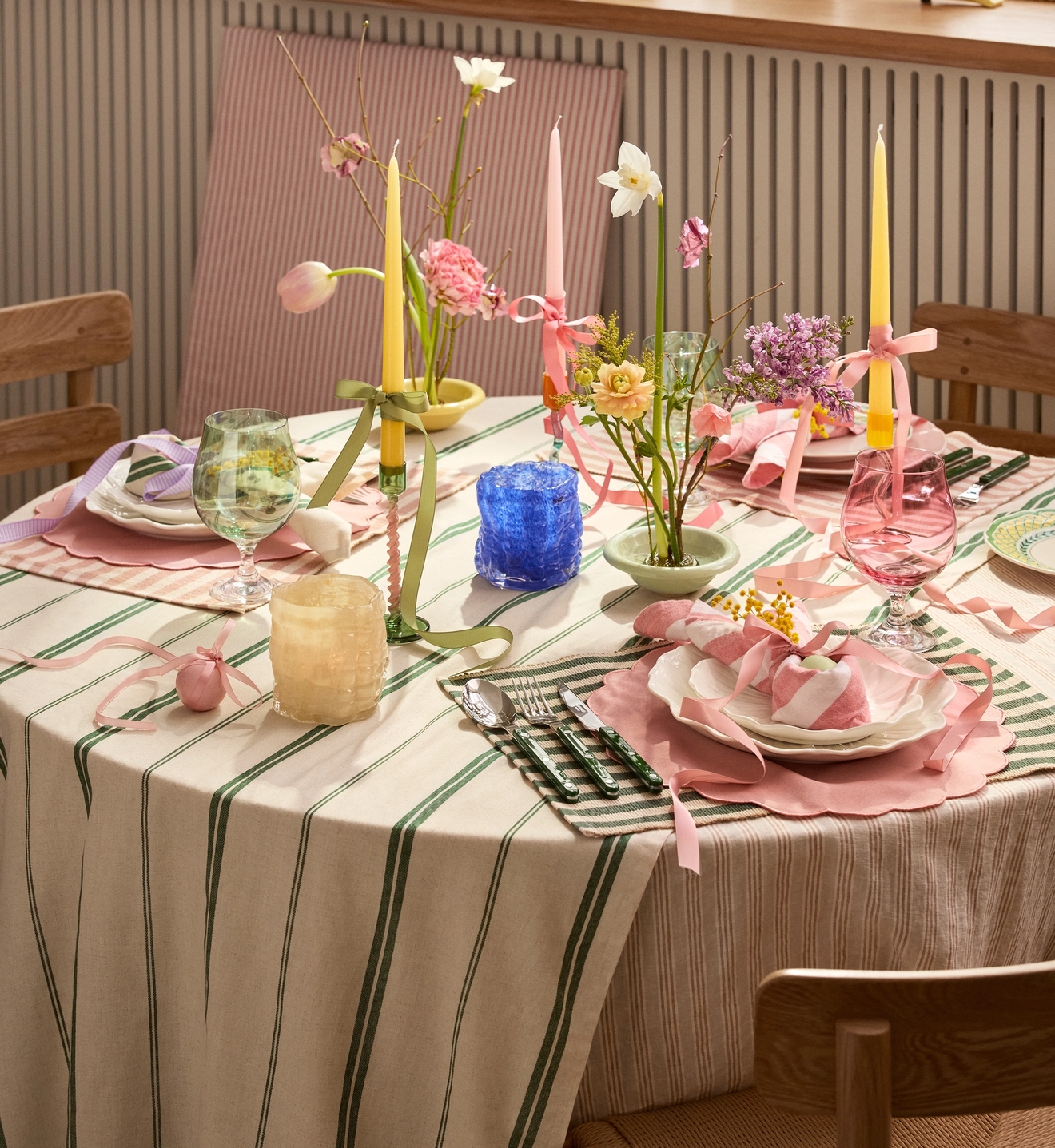 A festive spring dining table set with a green striped tablecloth, pink placemats, colorful candles, and bouquets of spring flowers.