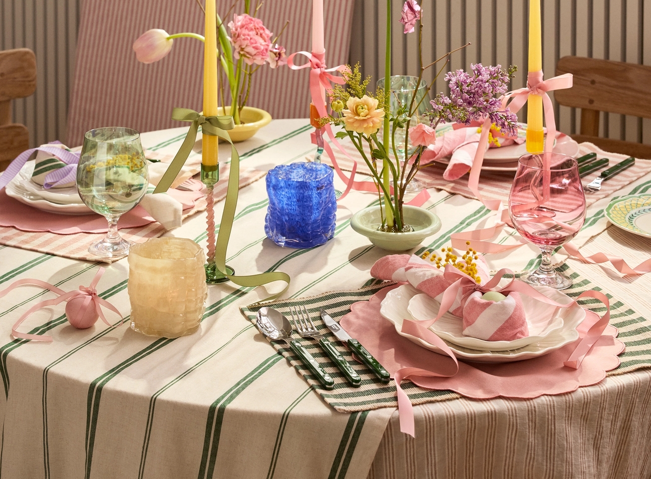 A vibrant spring table setting with a striped tablecloth, pink placemats, colorful candles, and bouquets of spring flowers.