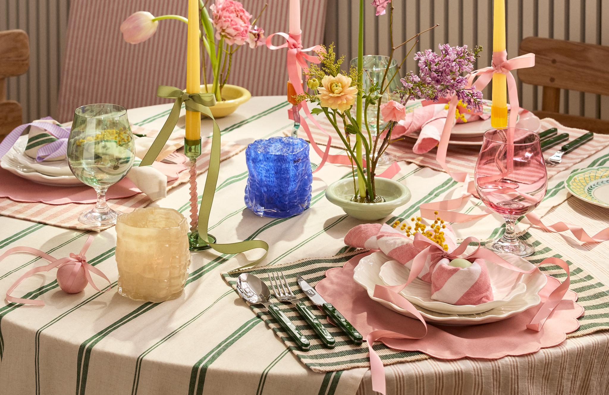 Kauniisti katettu kevätpöytä raidallisella pöytäliinalla, vaaleanpunaisilla tableteilla, kukka-asetelmilla ja pastellivärisillä kynttilöillä.