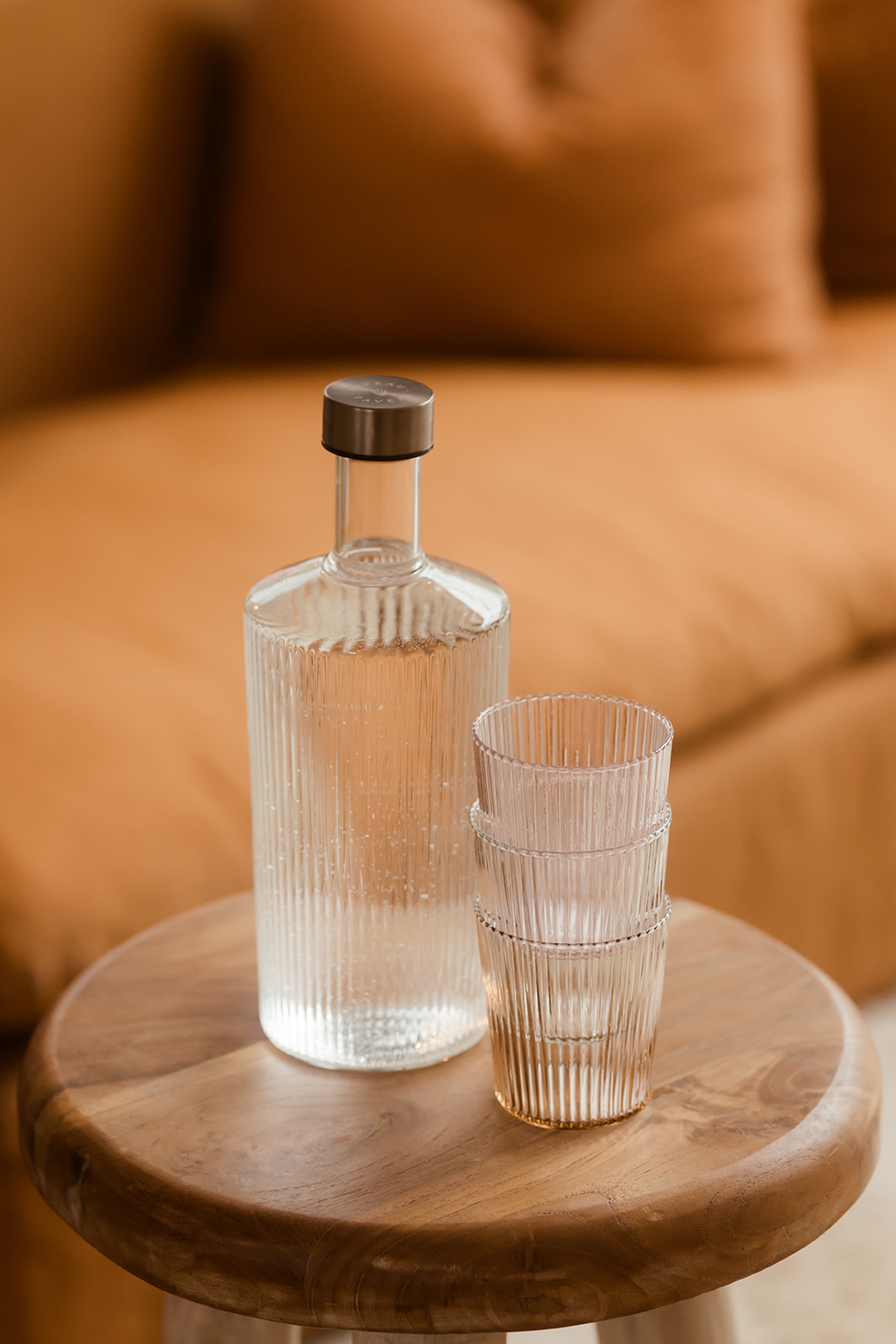 En räfflad karaff och räfflade glas i klart glas som står på ett litet soffbord framför en orange soffa